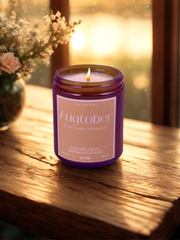 Candle in a amber glass jar with a beige rectangle label on the front. the candle is sitting on a medium toned wood surface. there is a vase of flowers off to the left and a out of focus window with light shining through the background.