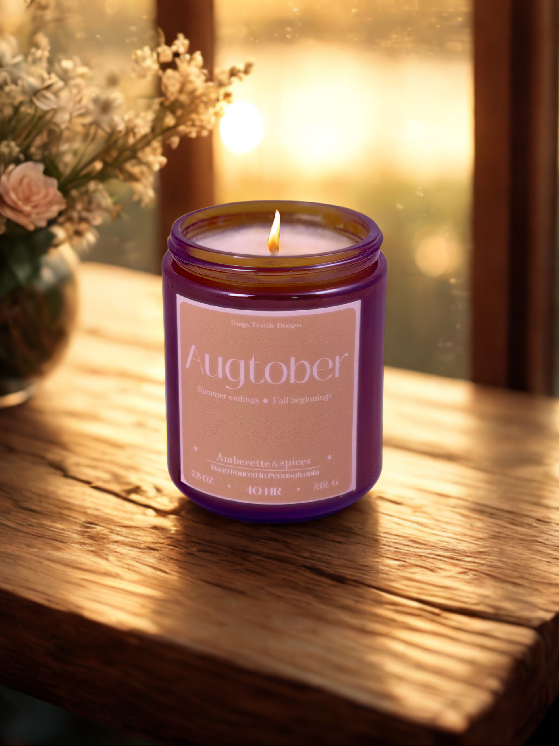 Candle in a amber glass jar with a beige rectangle label on the front. the candle is sitting on a medium toned wood surface. there is a vase of flowers off to the left and a out of focus window with light shining through the background.