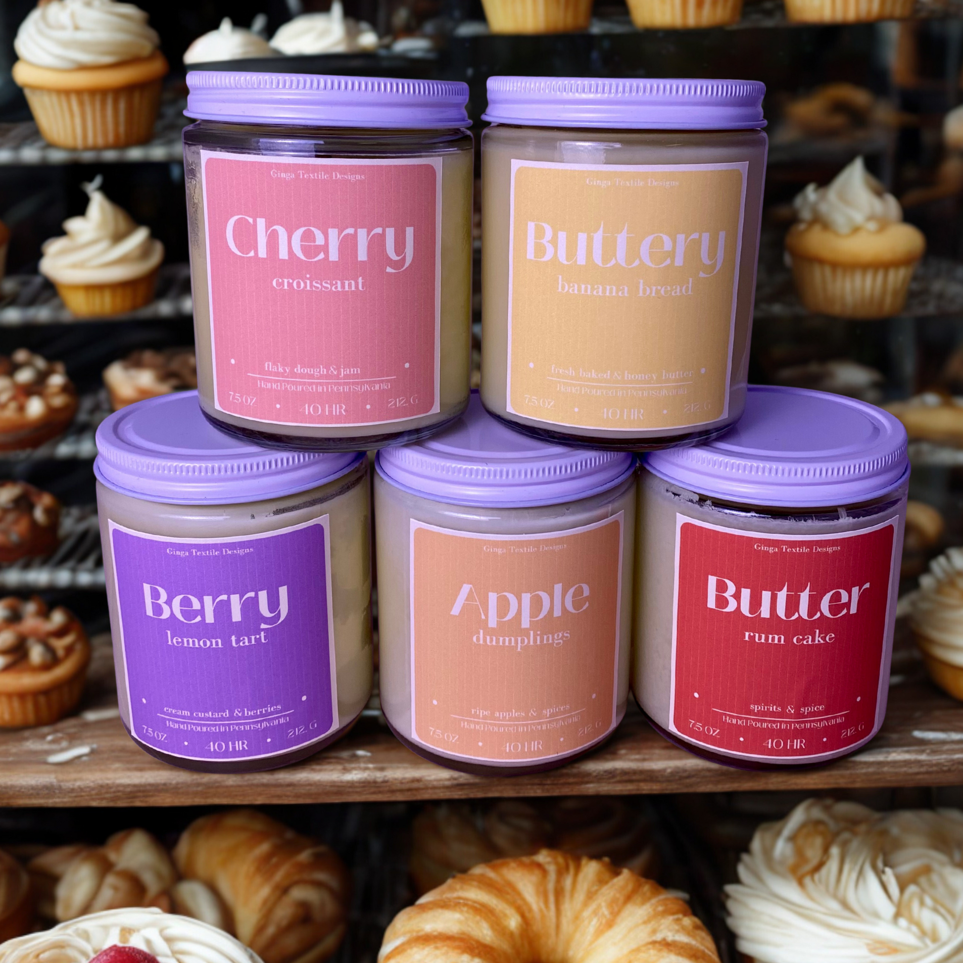 Five scented candles from the bakery collection with white lids and labels in front of a display of pastries.