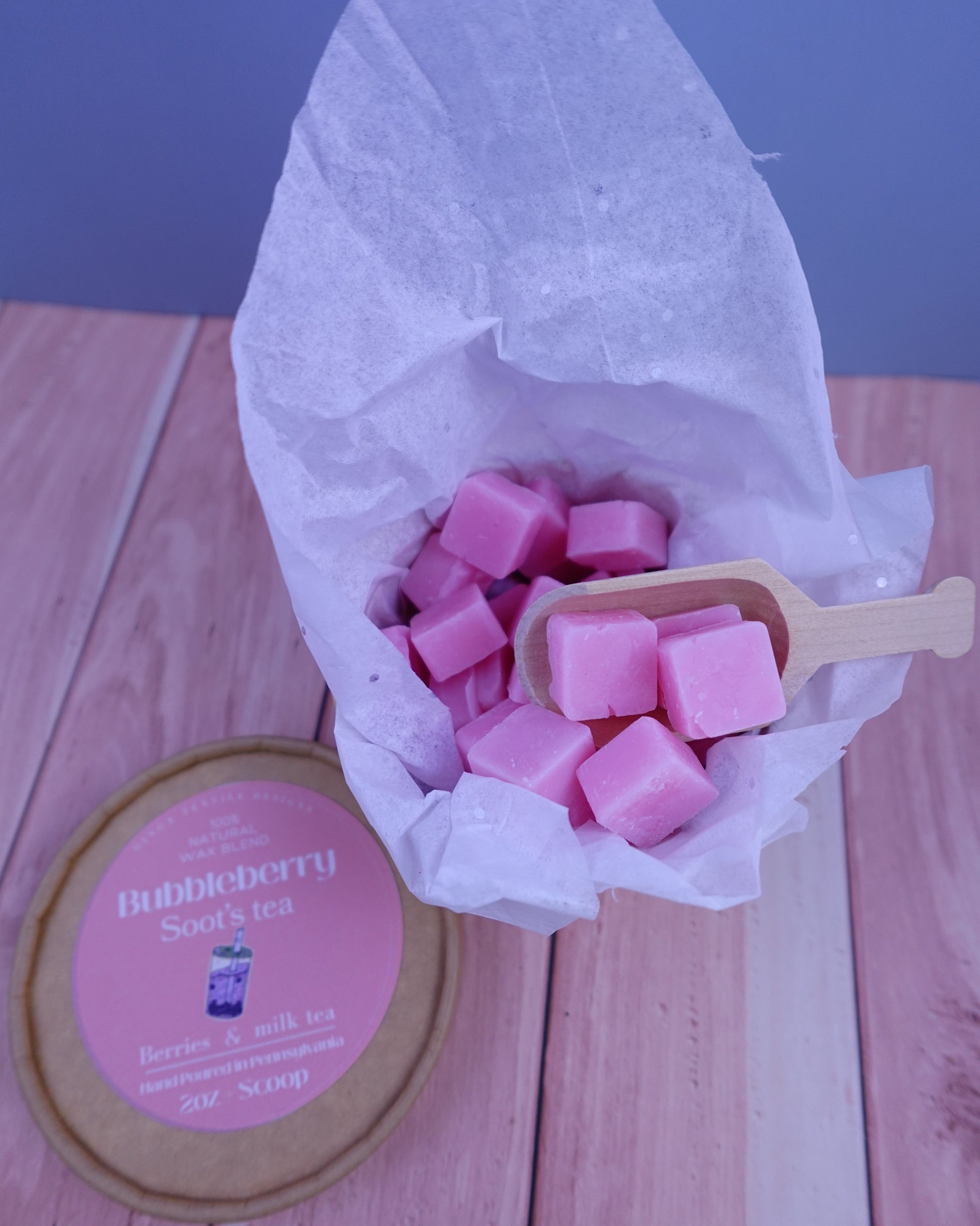 Pink wax cubes in a wooden scoop with a pink container labeled 'Bubbleberry soot's tea' on a wooden surface.