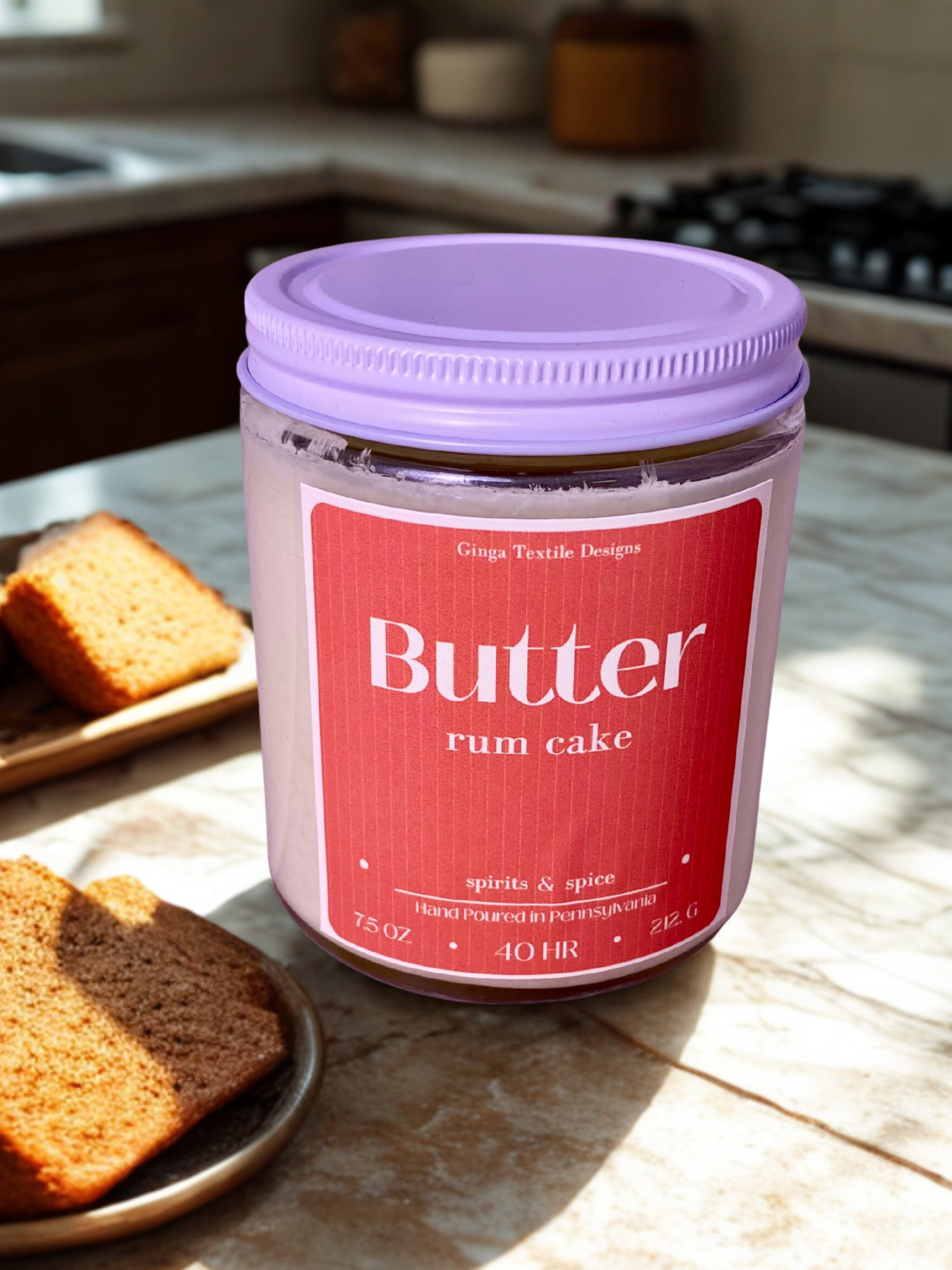 scented candle of 'Butter Rum Cake' on a kitchen counter with cake slices nearby and a blurred out of focus background of a kitchen 