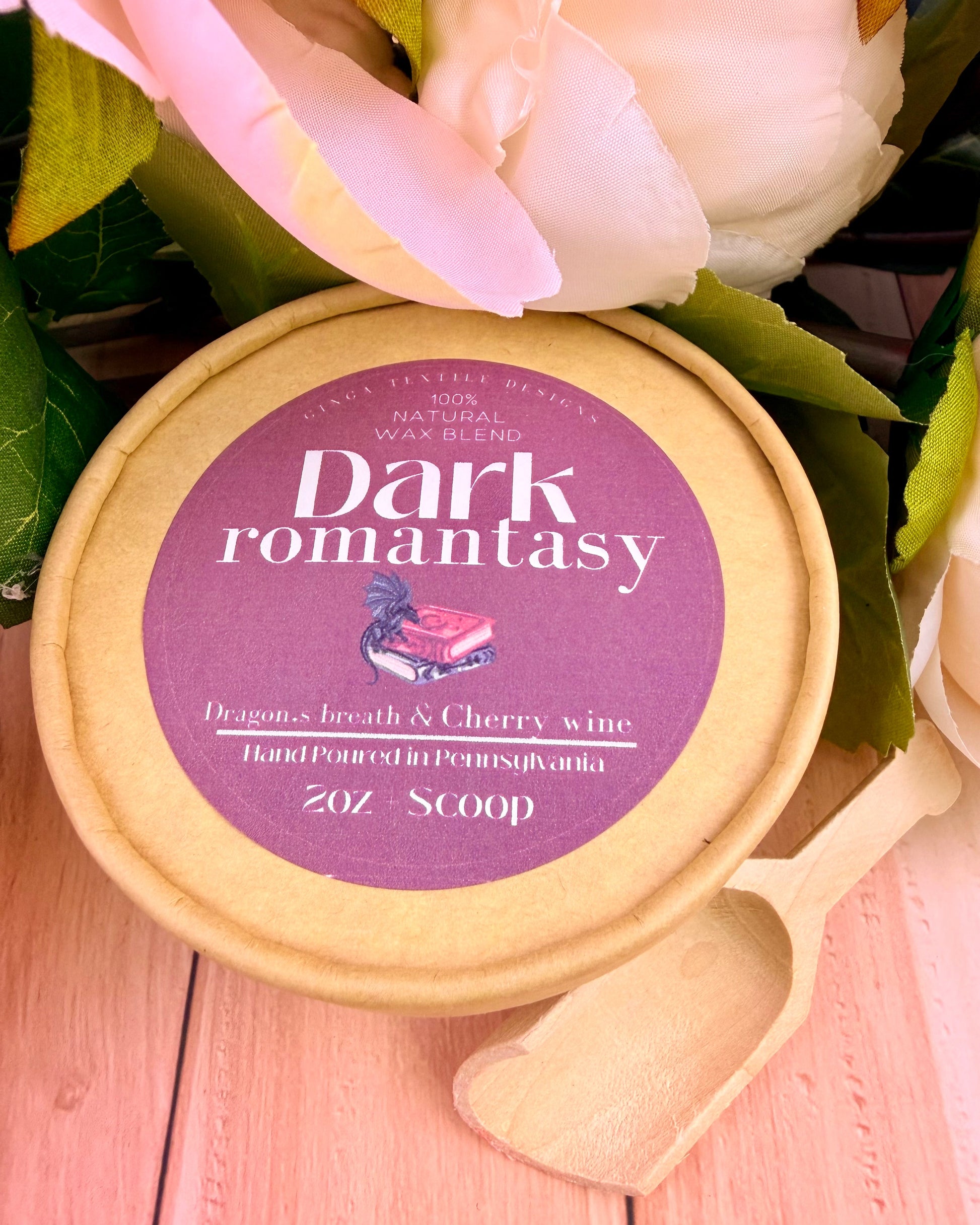 Packaging of wax melts of a scent called Dark Romantasy with a wooden scoop sitting on a light-colored wood table and peony flowers in the background