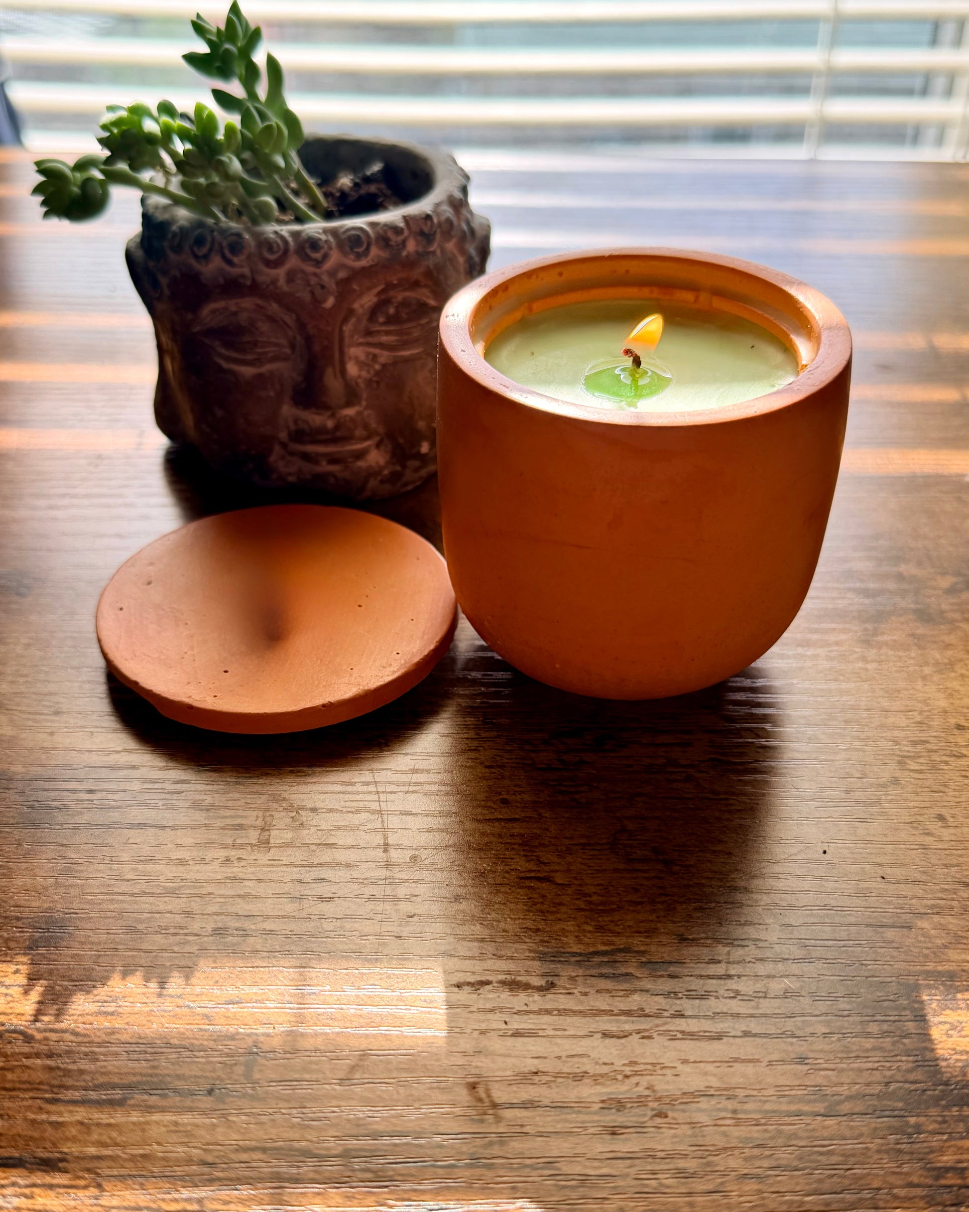 Ceramic pot with a candle and lid on a wooden surface, next to a small potted plant.