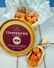 Heart shaped Wax tarts labeled 'Thumbprint cookies' on a wooden surface in a cardboard container with a wooden scoop and blue in the background.