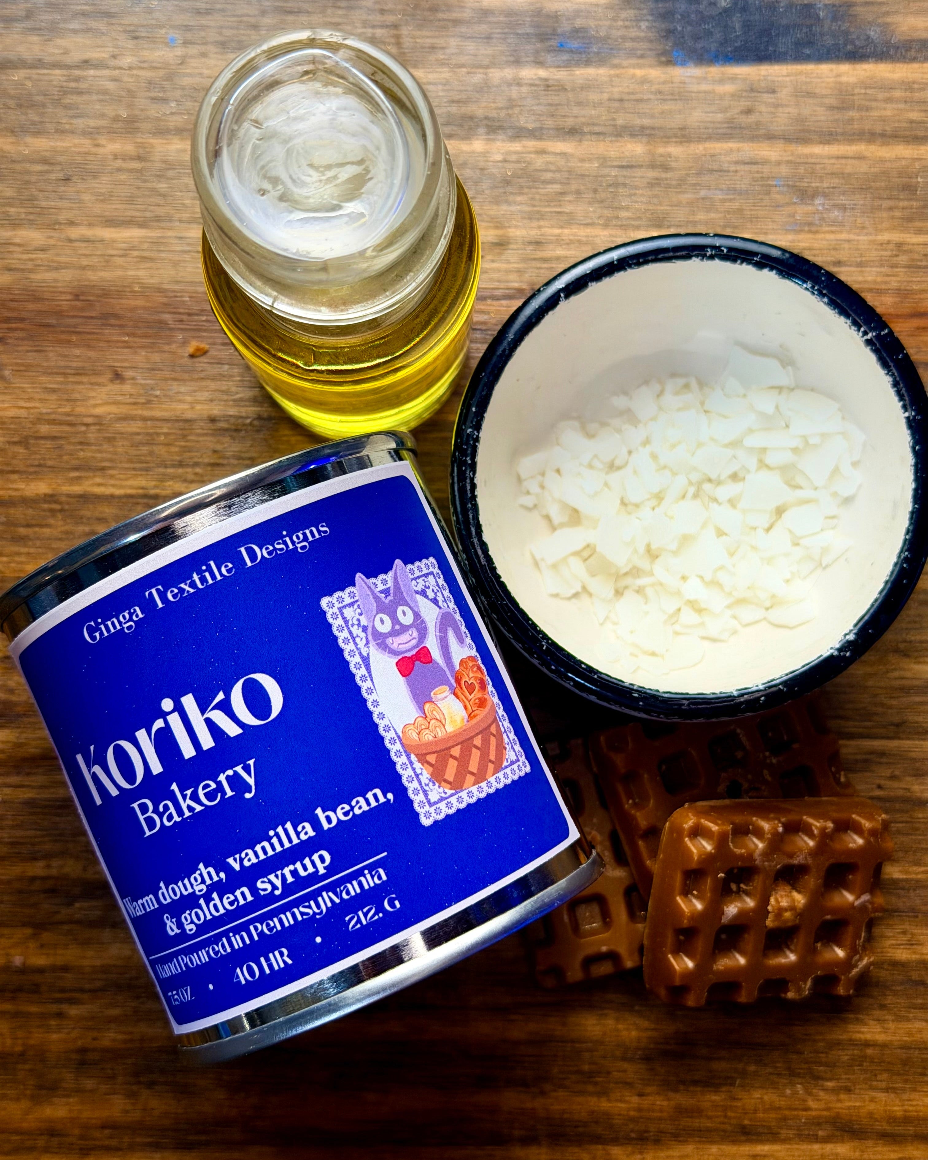 Container of Koriko Bakery product with a small bowl of white wax substance and waffle wax melt on a wooden surface and glass jar with golden syrup