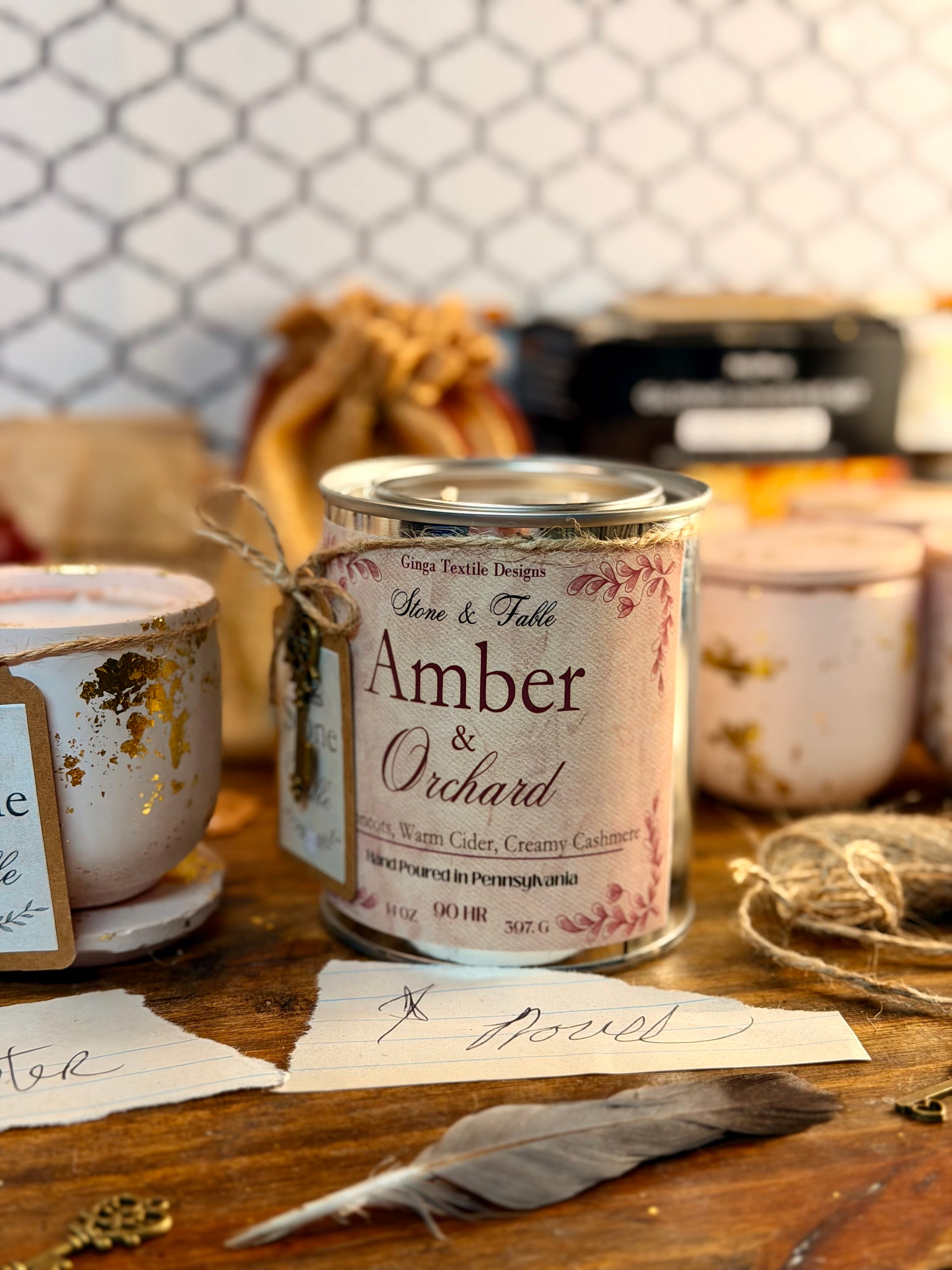 A metal candle tin labeled 'Amber & Orchard' by Stone & Fable Designs sits on a rustic wooden table with twine, paper tags, and other candles in the background.