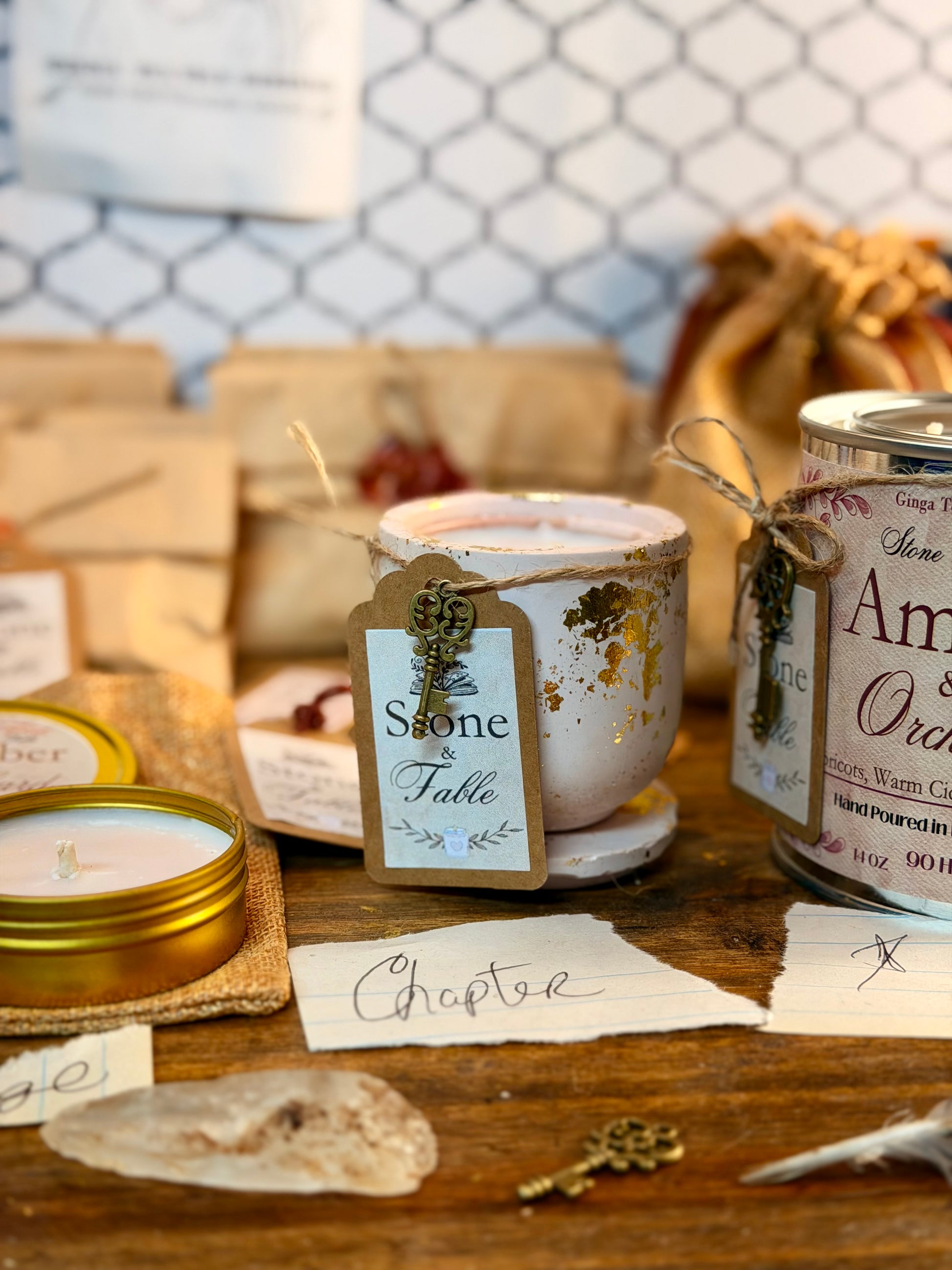 A styled tabletop display of candles and gift-wrapped packages on a wooden surface, with a central cream candle jar tied with twine and a paper tag reading 'Stone on Fable,' alongside other wrapped items and labels in a rustic setting.