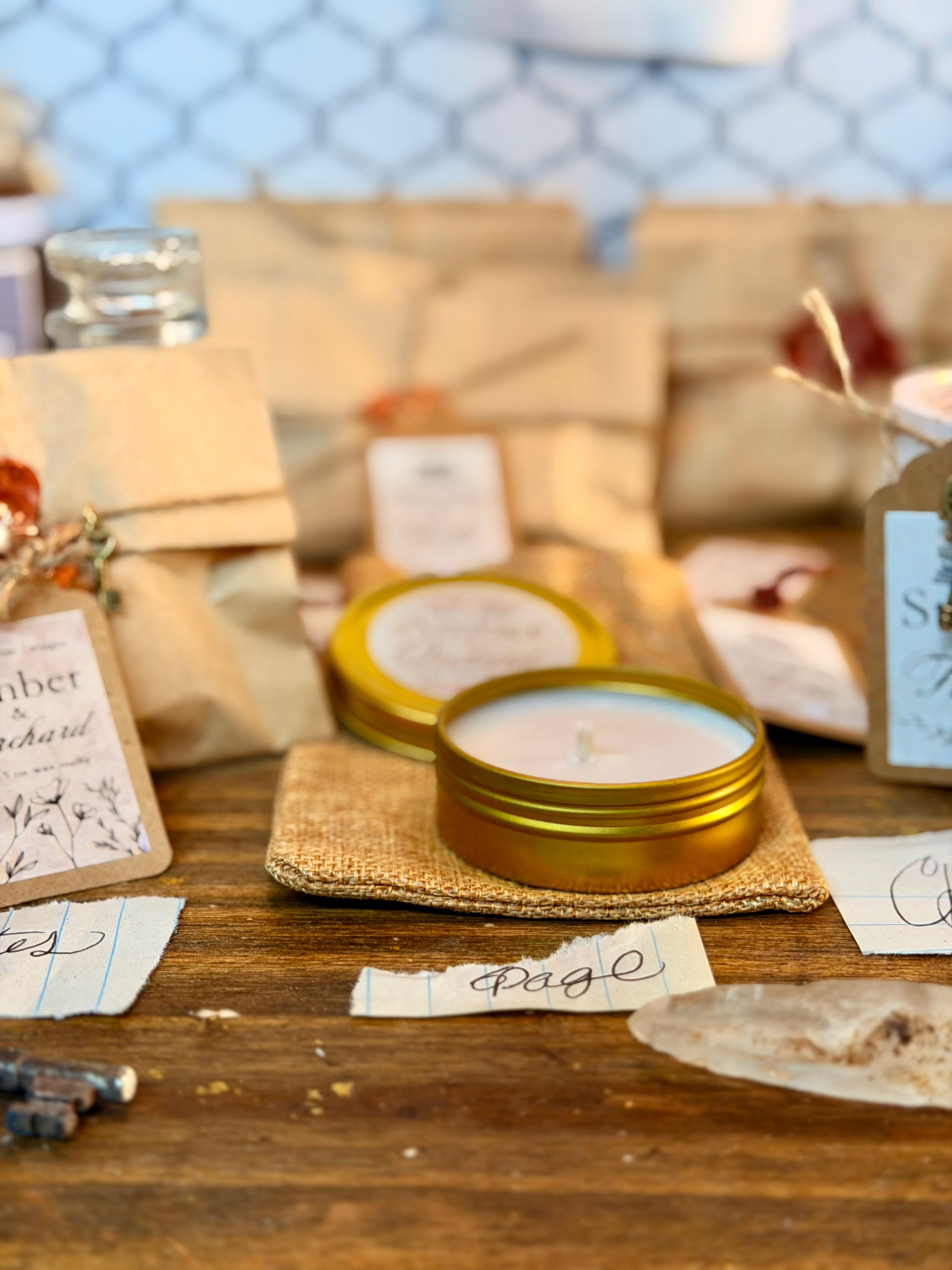 A small round metal tin candle with a gold lid sits on a burlap pad on a wooden table, surrounded by wrapped packages, tags, and jars in a styled, rustic setting.