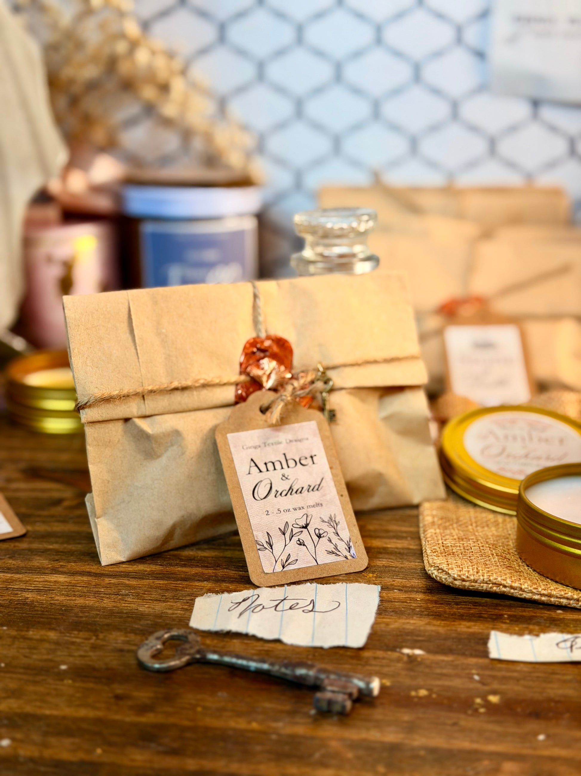 A styled product display featuring a kraft paper envelope tied with twine and a paper tag reading 'Amber Orchard', alongside candles in gold tins, a small glass bottle, and rustic paper notes on a wooden tabletop.