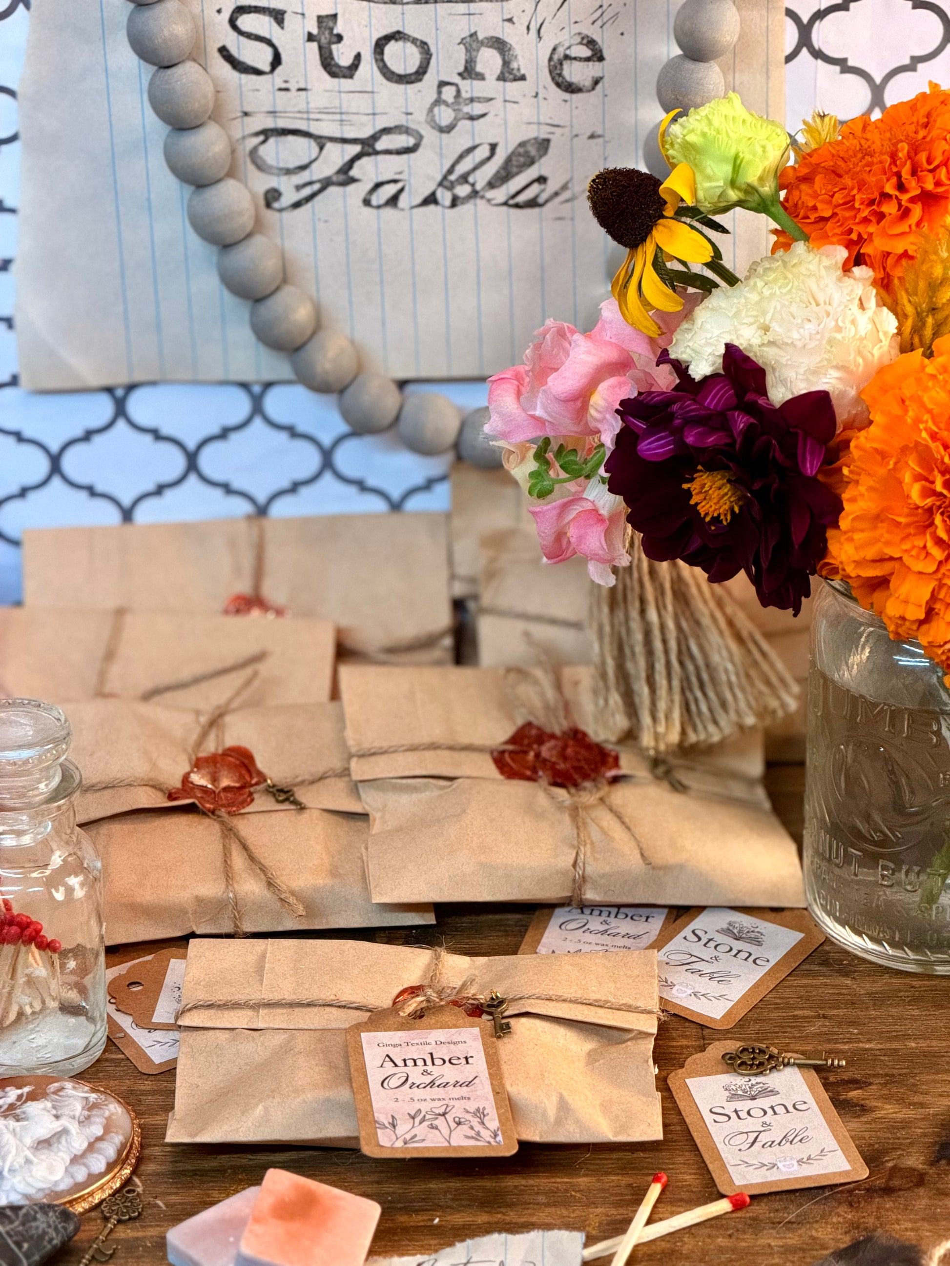A styled tabletop display featuring multiple small packages wrapped in kraft paper with twine and wax seals, several printed tags reading 'Amber & Orchard' and 'The Stone & Sage', a glass jar, dried flowers, and a colorful flower arrangement in a mason jar. A decorative sign in the background reads 'Stone & Fable'.