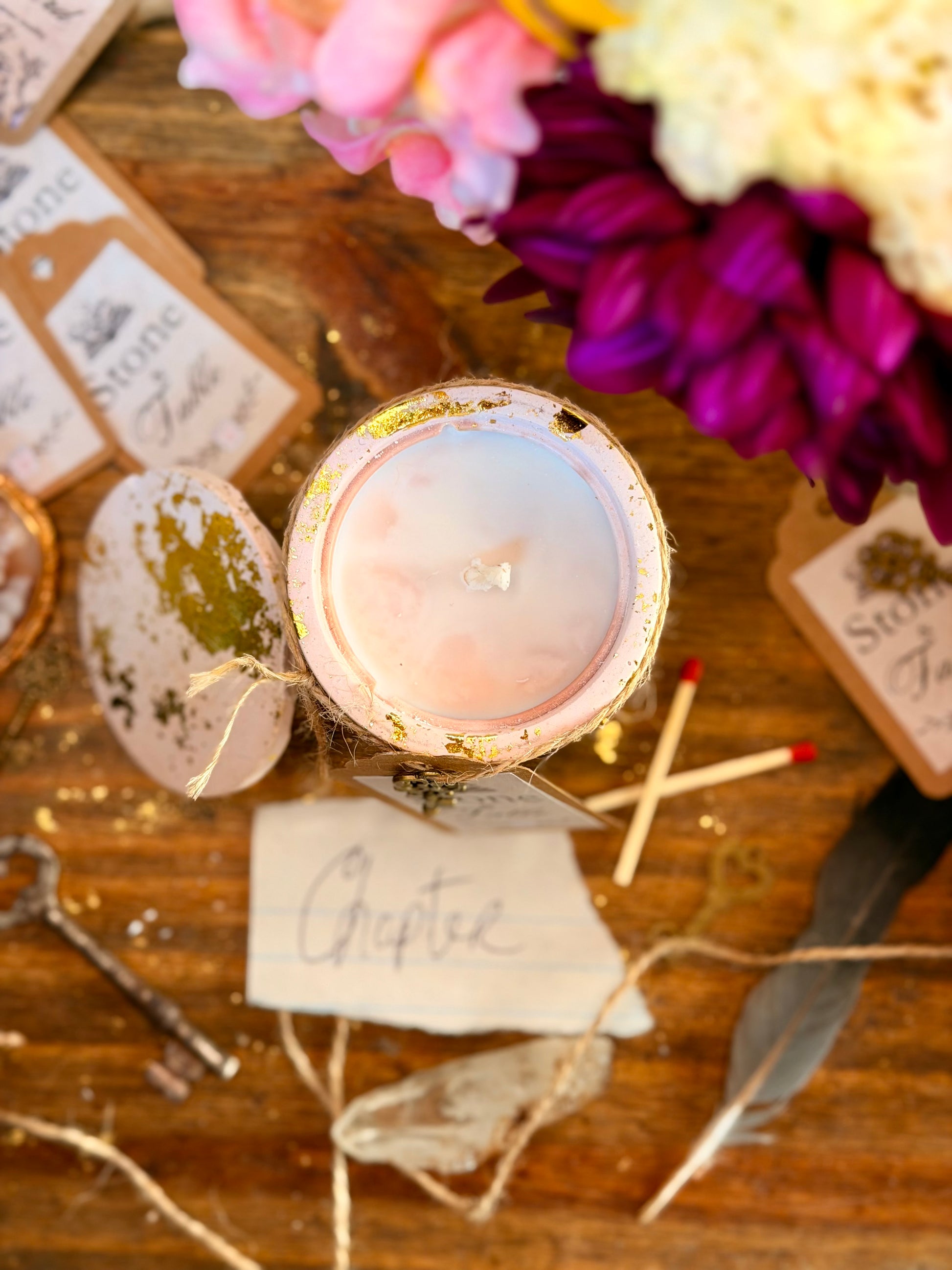A styled flat-lay of a small glass jar candle with a pale pink wax surface and a gold glitter rim, surrounded by rustic props including flowers, matchsticks, tags, twine, and a feather on a wooden surface.