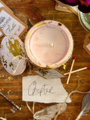 A styled flat-lay of a candle in a distressed pink ceramic jar with gold leaf accents, shown alongside decorative props on a wooden surface.