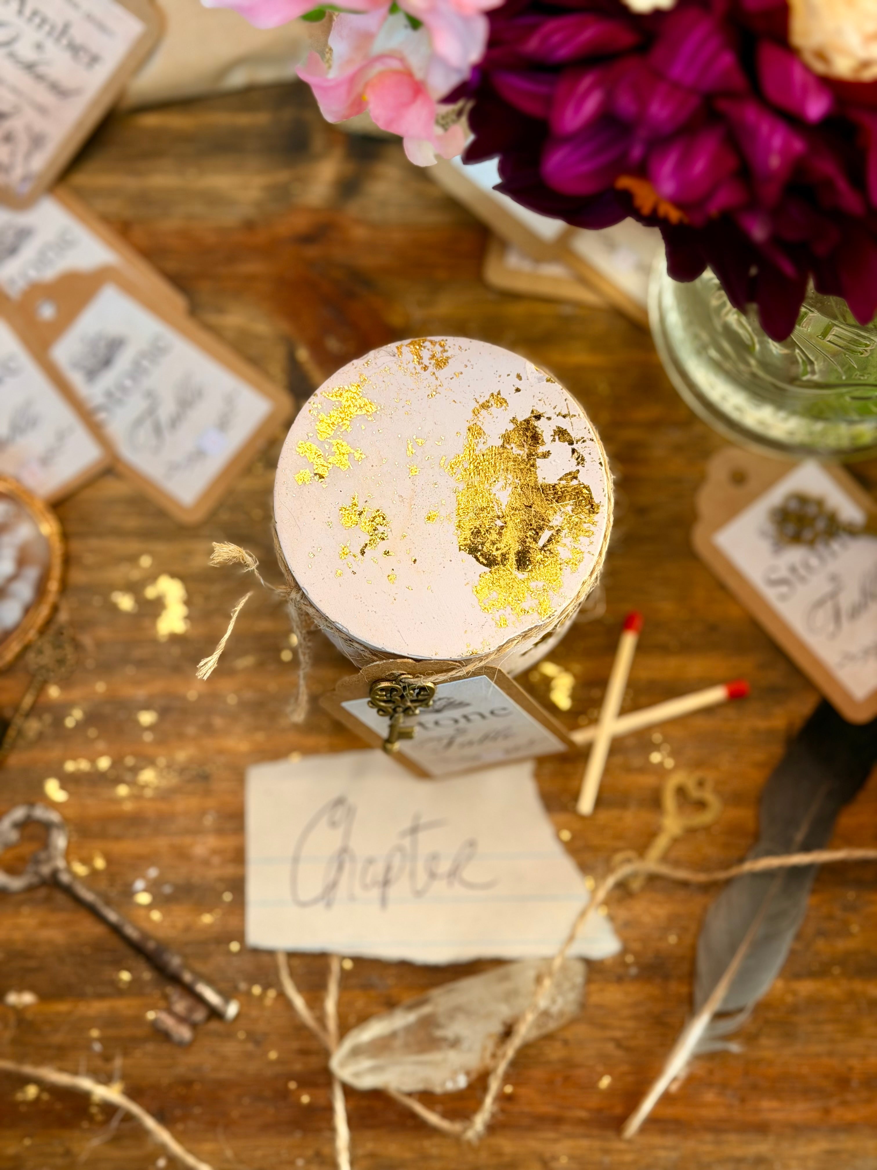 A small round cement jar with a white lid decorated with gold foil flakes, tied with twine and a small tag, displayed on a rustic wooden table surrounded by wedding-themed stationery, flowers, keys, and matchsticks.