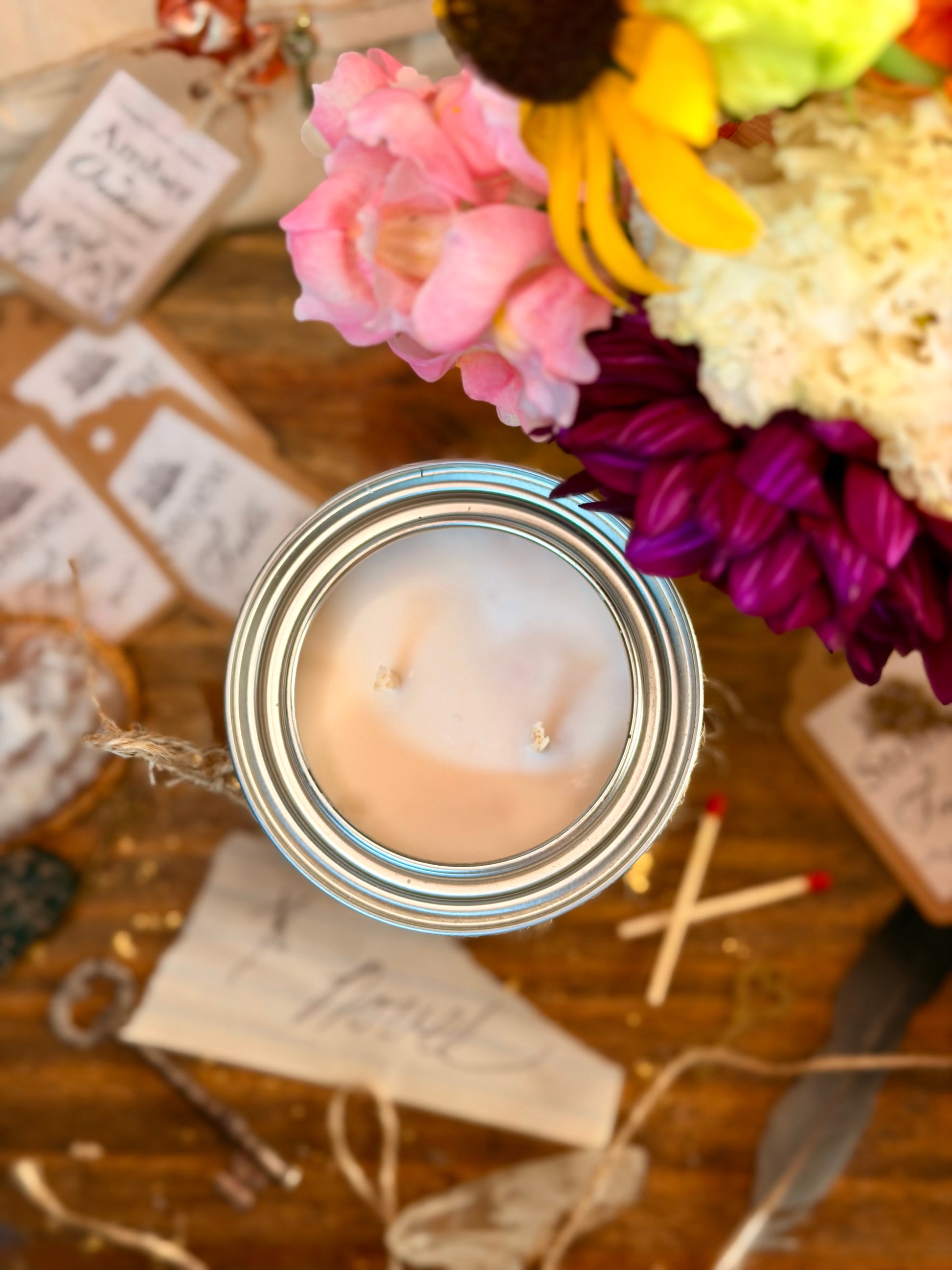 A top-down view of a double-wick candle in a silver metal jar, surrounded by rustic decorative props including flowers, matchsticks, paper tags, and twine on a wooden surface.