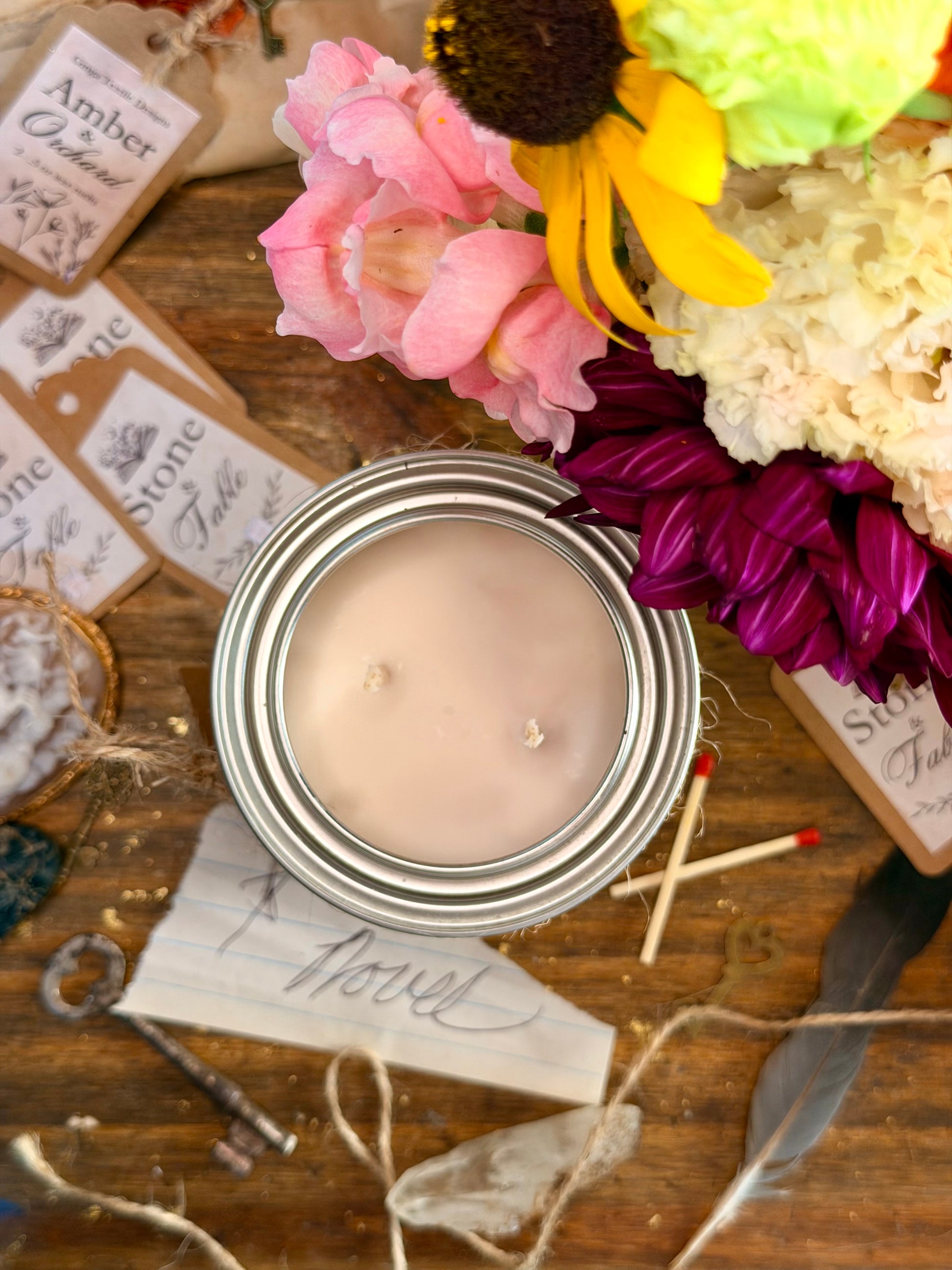 An open metal candle jar filled with pale blush pink wax, styled on a rustic wooden surface with flowers, paper tags, twine, a key, and small matches.