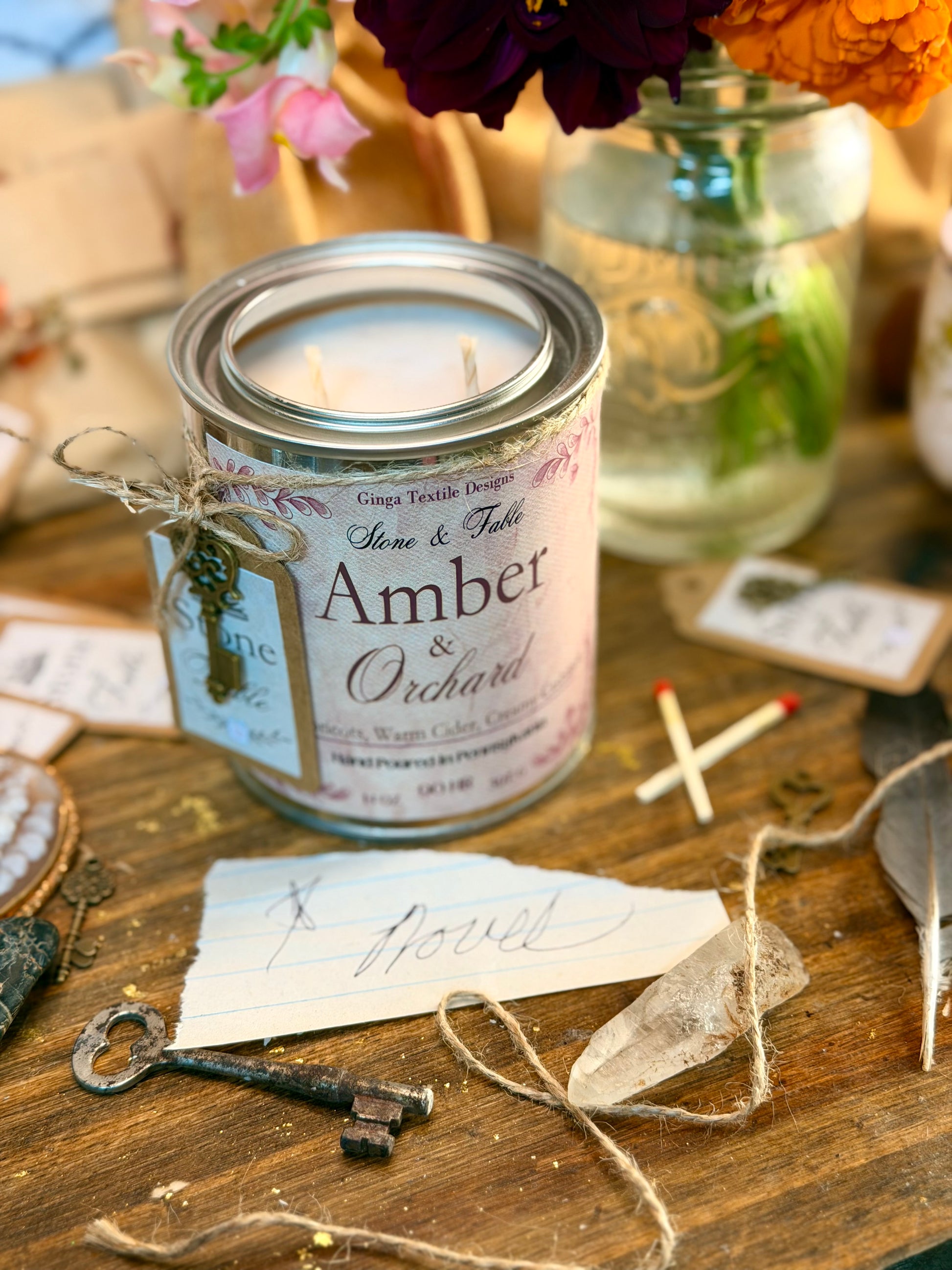 A tin candle labeled 'Amber & Orchard' by Ginga Textile Design, styled on a rustic wooden table with twine, a vintage key, paper tags, and floral elements in the background.