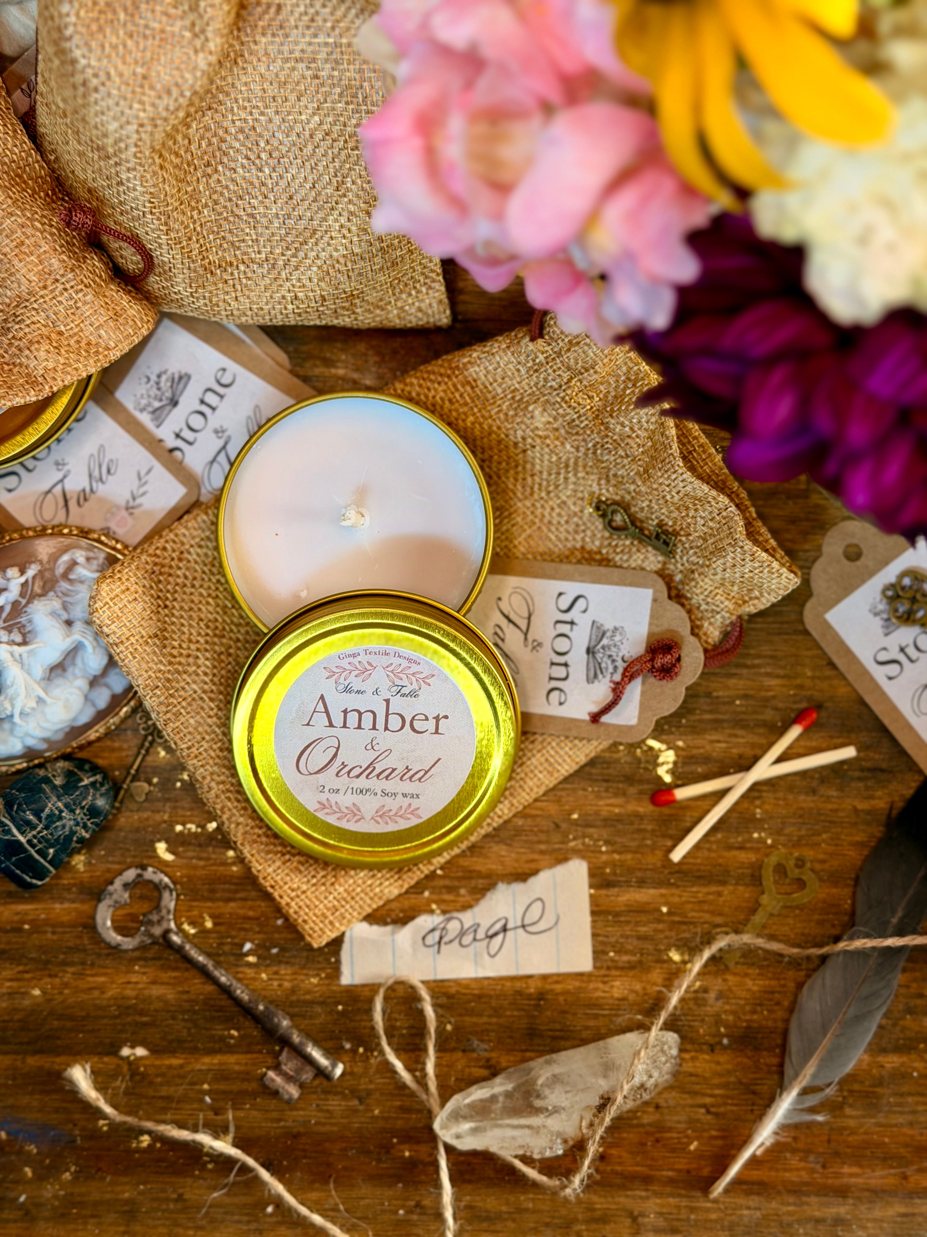 An open round tin candle labeled 'Amber Orchard' sits on a rustic wooden surface with burlap bags, flowers, tags, a key, twine, and small decorative props around it.
