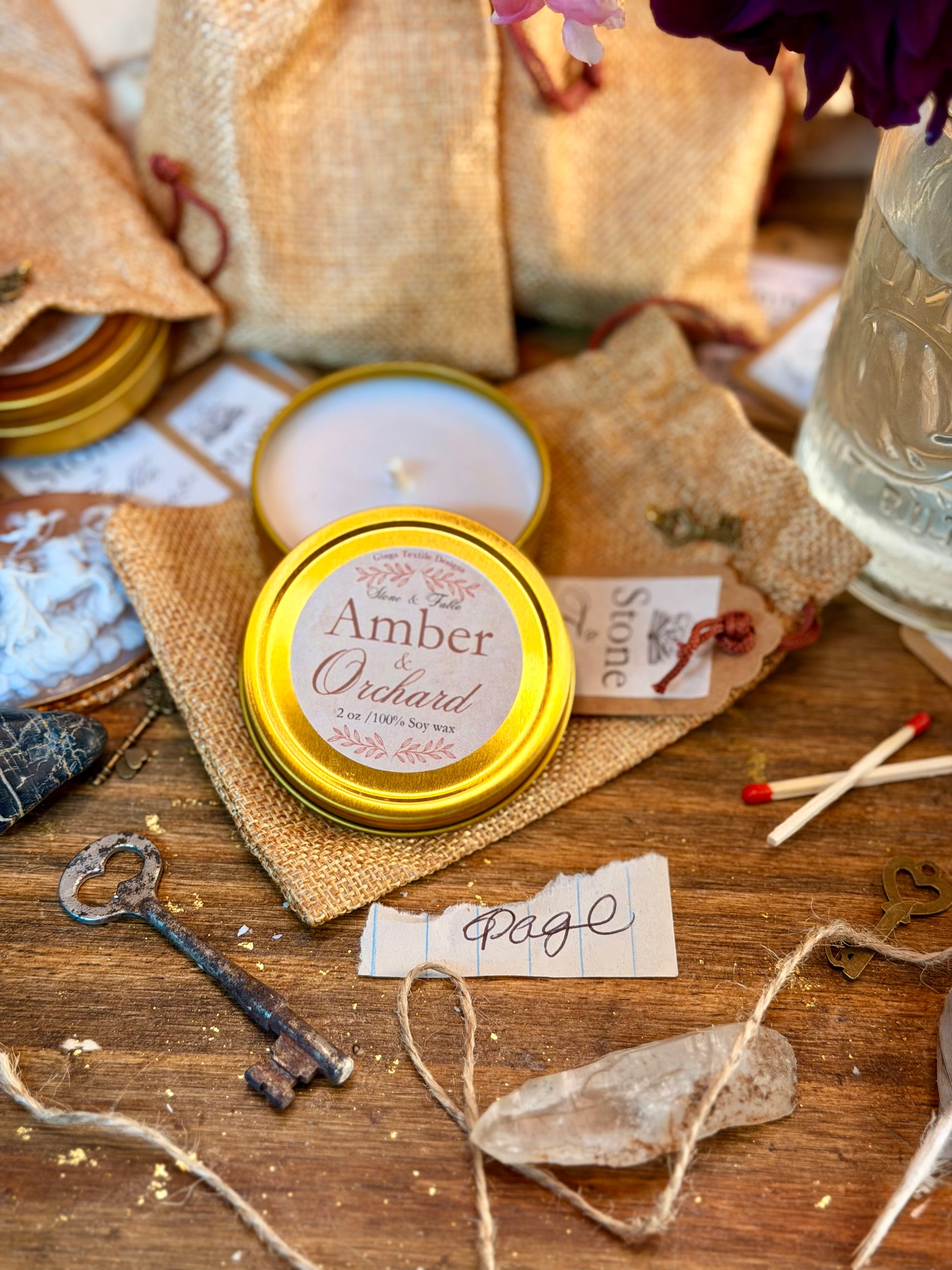 A round tin candle labeled 'Amber & Orchard' sits on a rustic wooden surface with burlap bags, a matchbook, keys, twine, and other styled props. A second candle is visible behind it, and the scene is arranged in a warm, handcrafted setting.