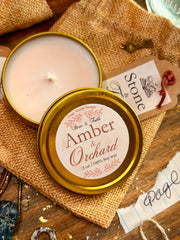 An open round candle tin showing pale cream wax, with the lid placed in front and printed label reading 'Amber & Orchard' and 'Stone & Fable.' The candle is styled on burlap fabric over a wooden surface with decorative keys, paper scraps, and a small red ribbon accent.