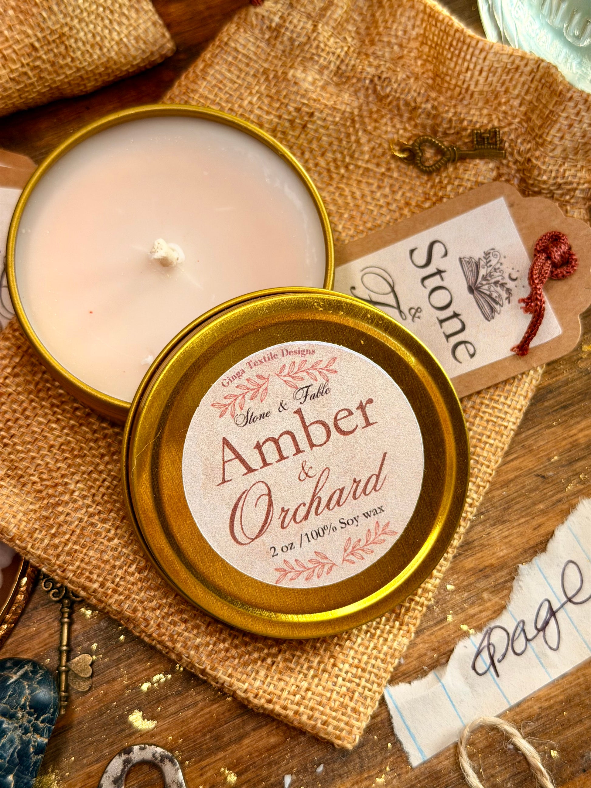 An open round candle tin showing pale cream wax, with the lid placed in front and printed label reading 'Amber & Orchard' and 'Stone & Fable.' The candle is styled on burlap fabric over a wooden surface with decorative keys, paper scraps, and a small red ribbon accent.