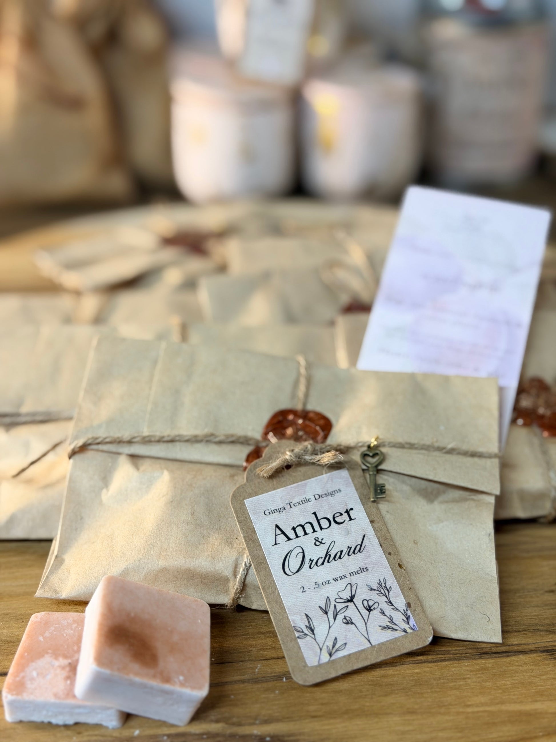 A close-up of packaged artisanal soap bars wrapped in kraft paper with twine and a wax seal, alongside a paper tag reading 'Amber & Orchard' and a small key. The scene is styled on a wooden surface with a softly blurred background of similar packages and jars.