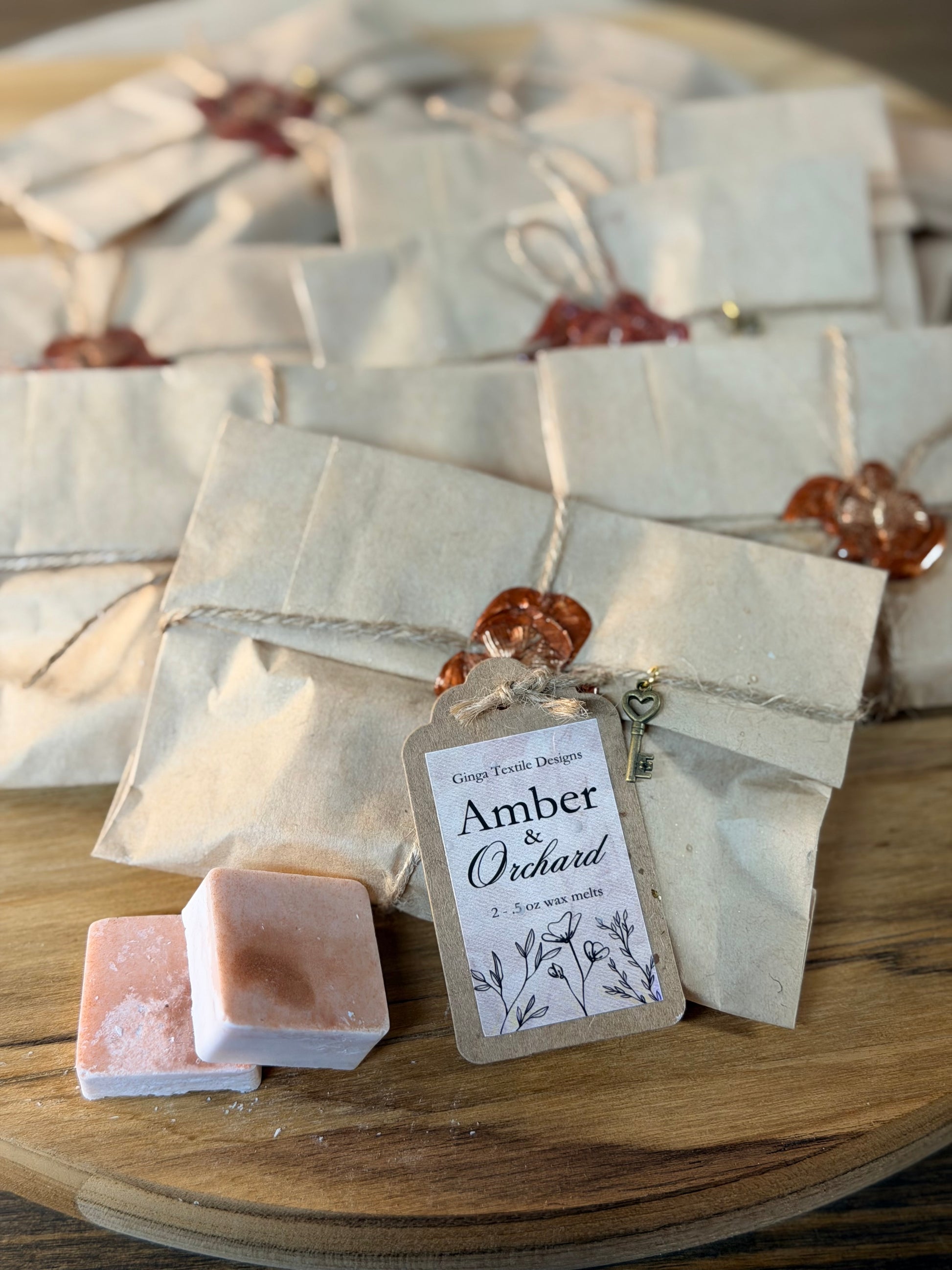 A styled product shot of handmade soap bars in pinkish-white packaging, tied with twine and sealed with amber wax seals, arranged on a wooden board. A small hanging tag reads 'Amber & Orchard'.