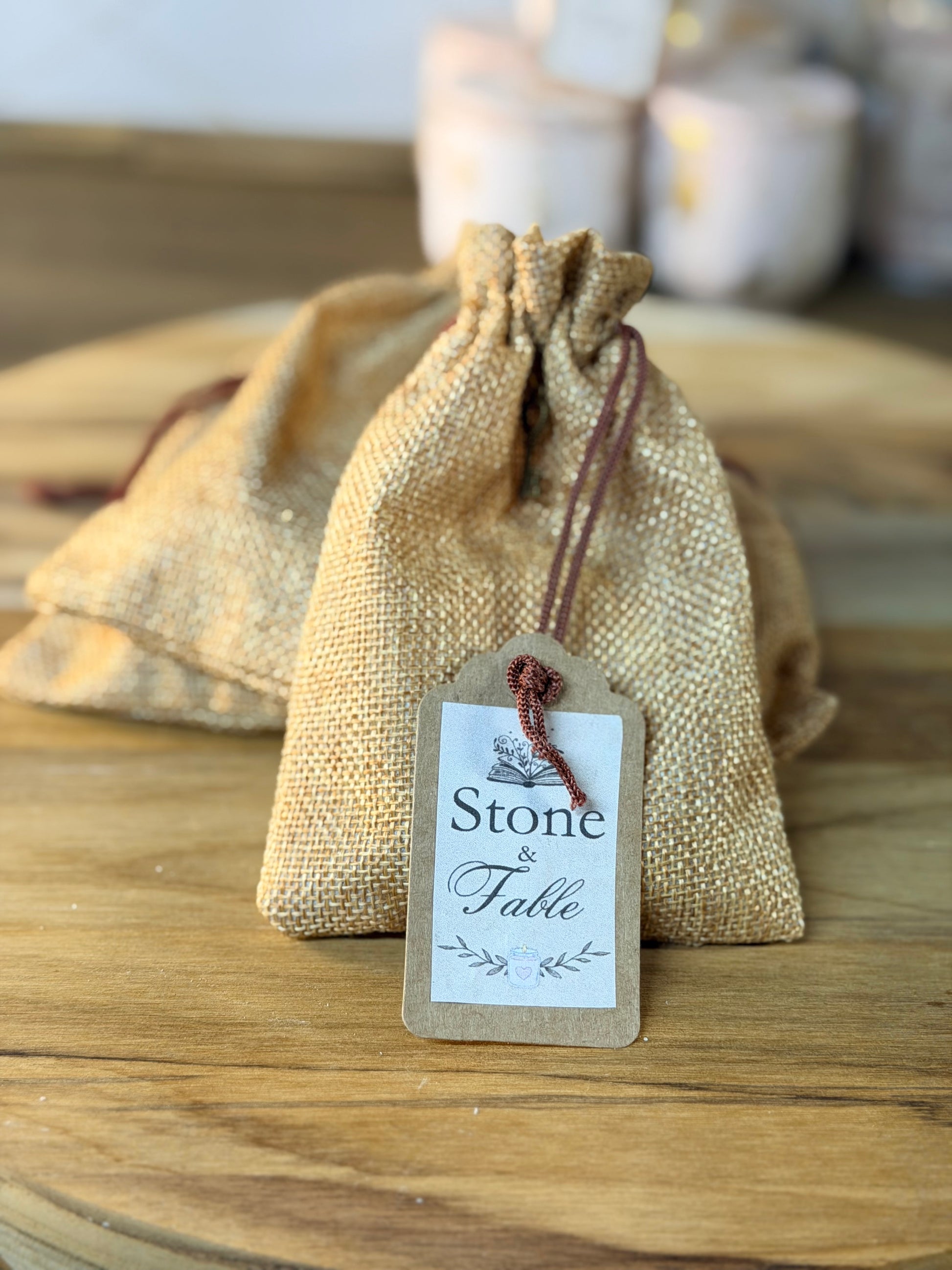 A small natural burlap drawstring pouch with a hanging paper tag reading 'Stone & Fable,' placed on a wooden surface with a softly blurred background.