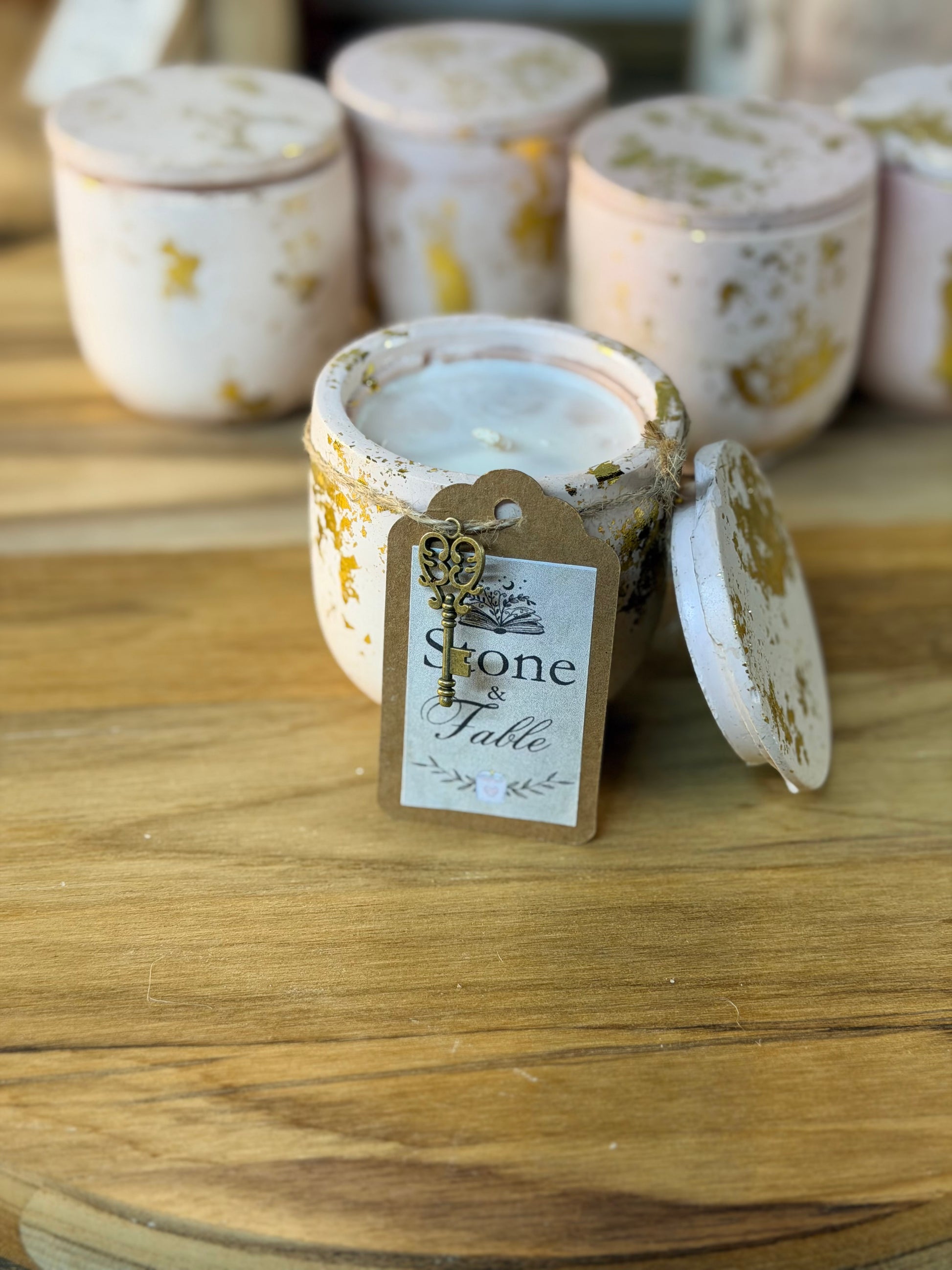 A white wax candle in a distressed, gold-leaf-patterned ceramic jar sits on a wooden table with its lid leaning against the front. A paper tag on the jar reads 'Stone Fable' with a decorative key charm.