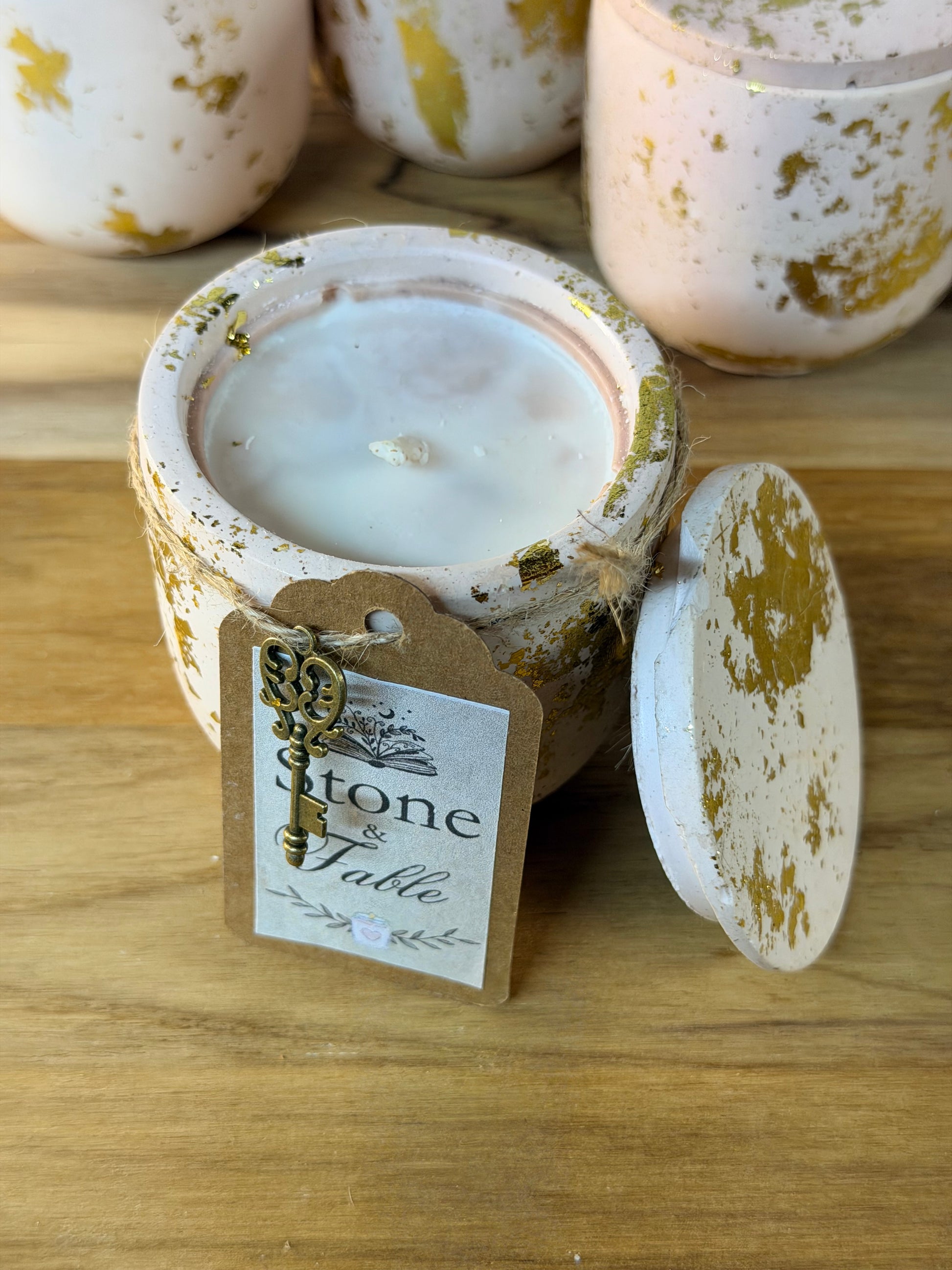 A ceramic candle jar with a white wax fill and distressed gold and beige glaze, shown open with its lid leaning against it on a wooden surface. A paper tag hangs from the rim and reads 'stone & Fable'.