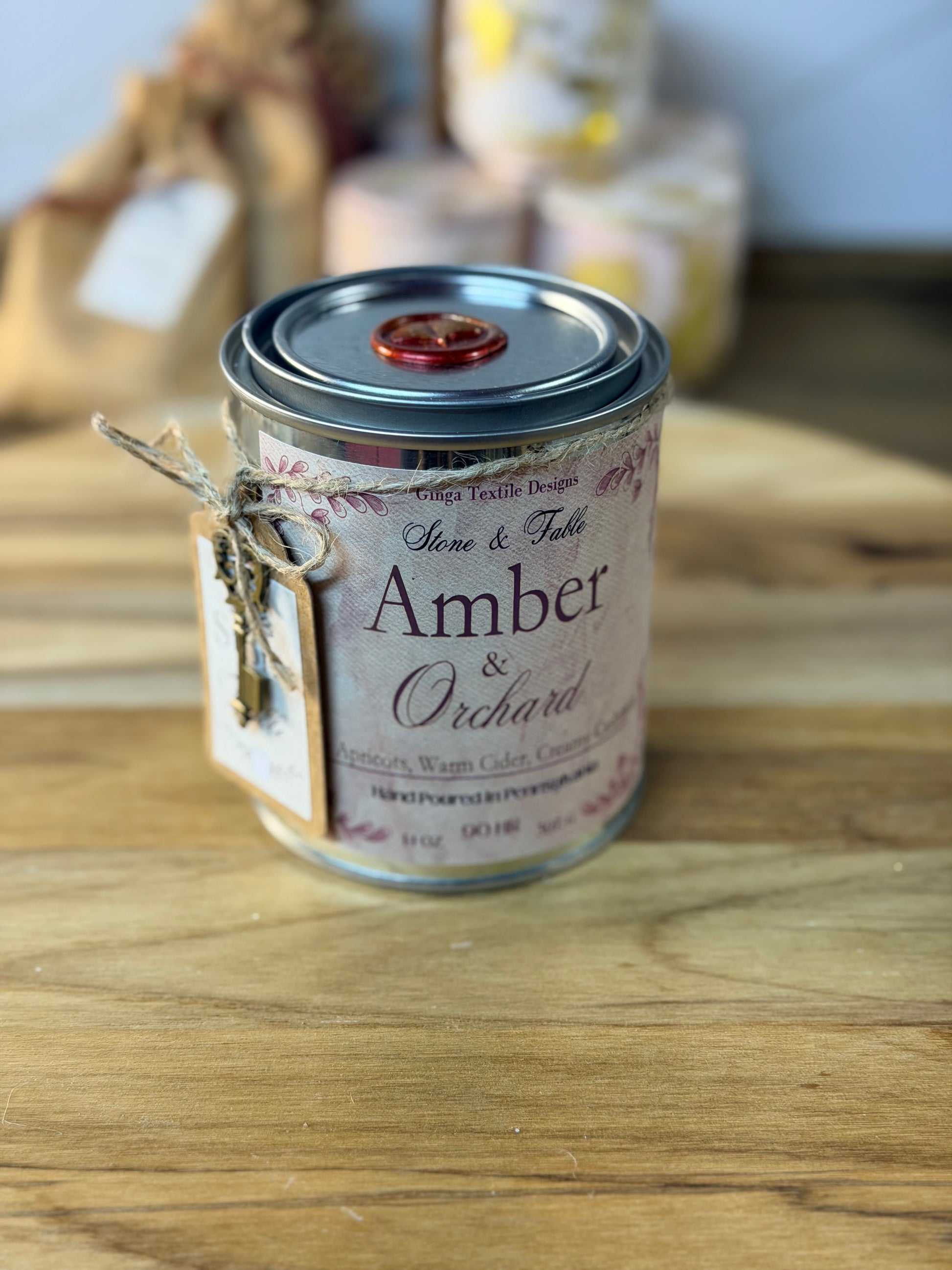 A small metal tin of Scents & Fabrics 'Amber & Orchard' scented wax melts sits on a wooden tabletop. The tin has a rustic kraft paper label tied with twine and a silver lid sealed with a red wax seal.
