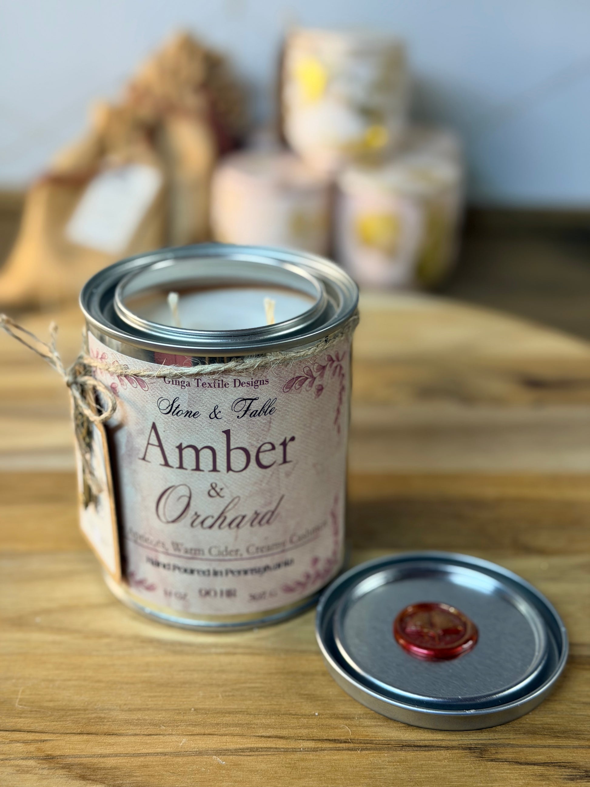 A metal tin candle labeled 'Amber & Orchard' sits on a wooden surface with its lid placed beside it. The candle has a beige label with pink floral script and a silver metal rim.