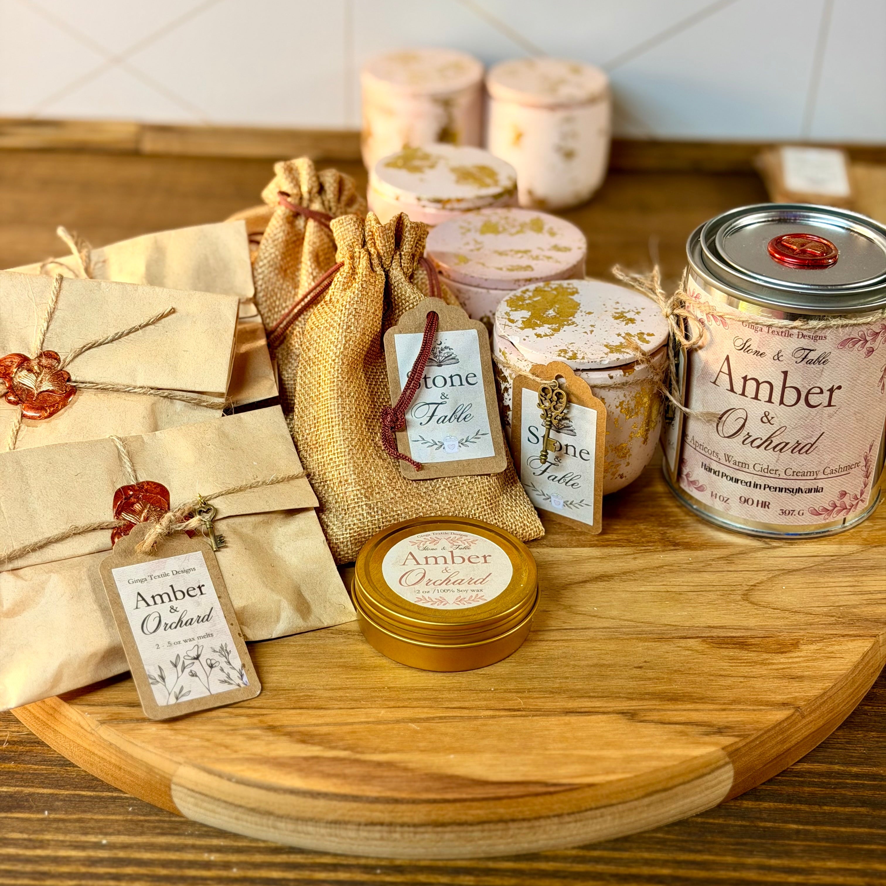 A styled product display of artisanal gift items on a round wooden board, including amber scented candles in jars, small wrapped packages with red wax seals, burlap drawstring bags, and tags reading 'Amber by Orchard'.