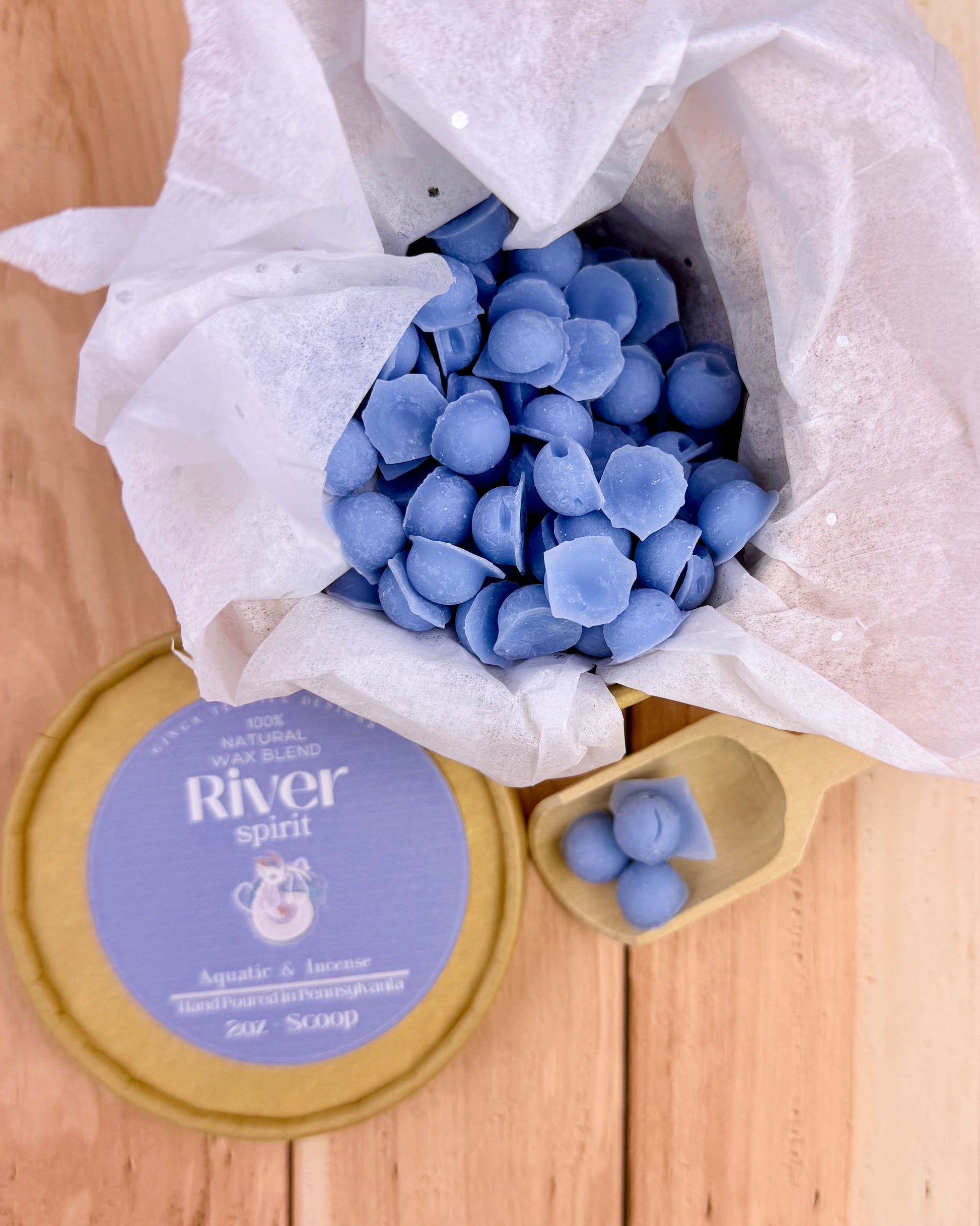 Blue wax melts in a cardboard container with a 'River Spirit' labeled lid on a wooden surface