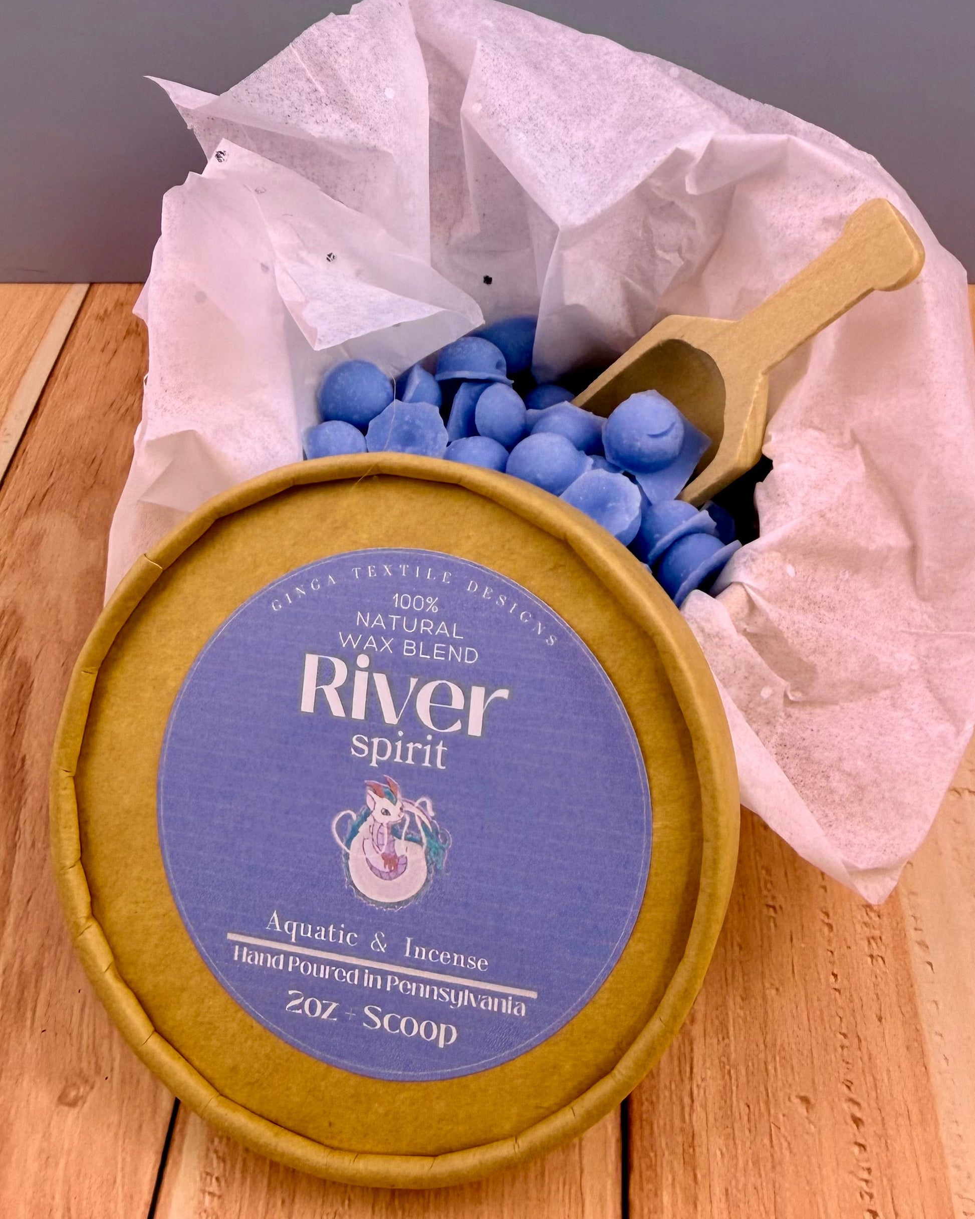 Container of blue wax beads with a wooden scoop and 'River Spirit' label on a wooden surface.