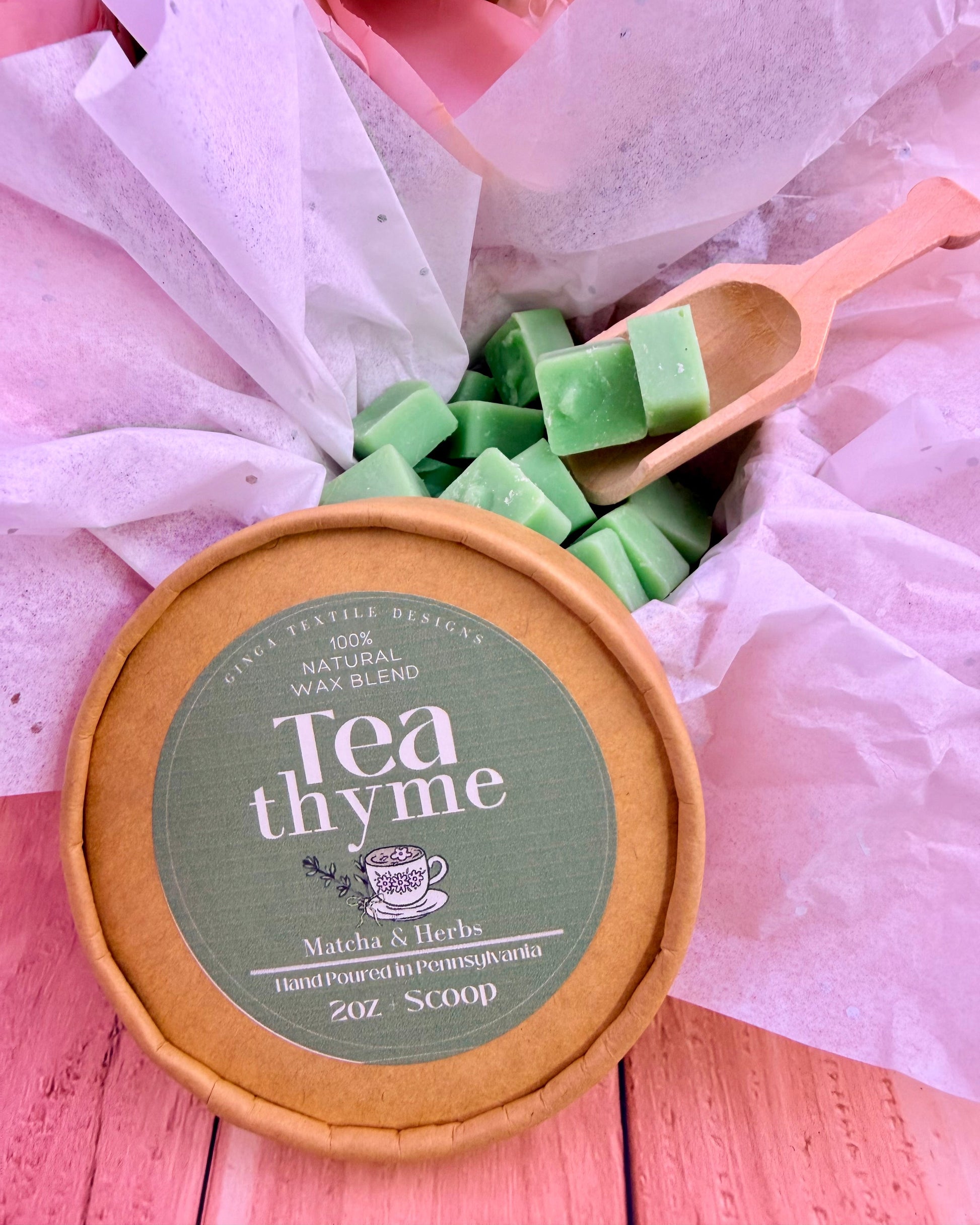 Container of 'Tea Thyme' wax melts with a wooden scoop on a pink surface.