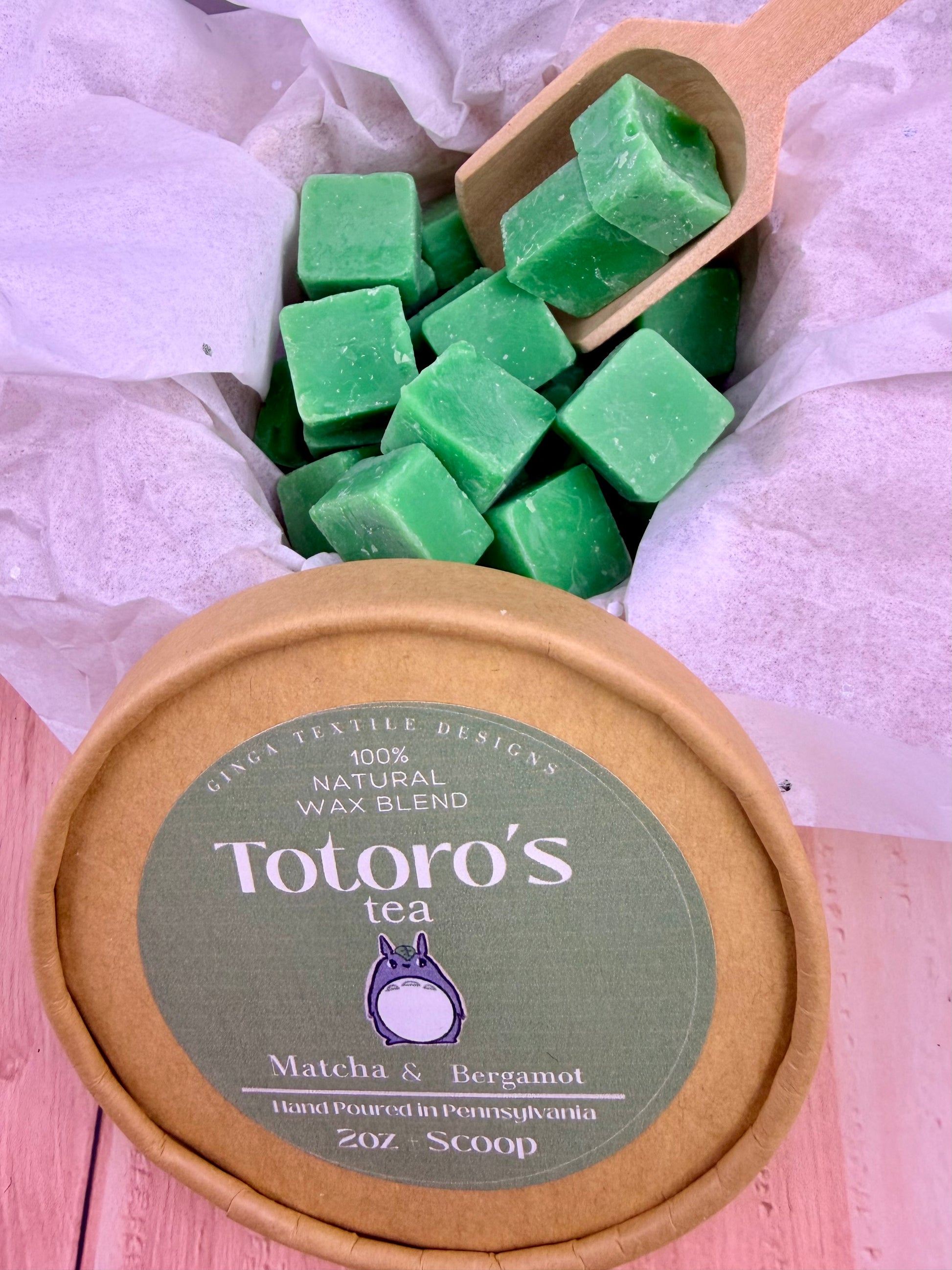 Green wax cubes in a wooden scoop with Totoro's Tea packaging on a pink surface