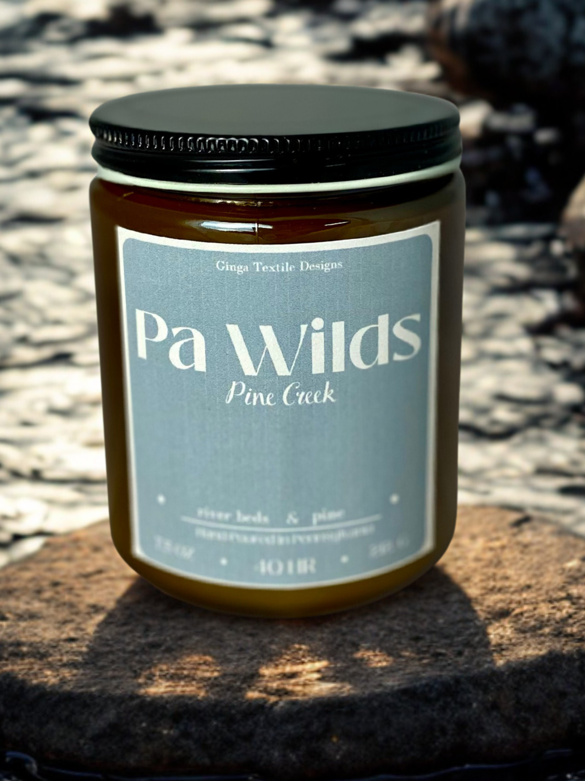 Amber glass scented candle in a jar with a black lid labeled 'Pa Wilds Pine Creek' on a stone surface with a natural background